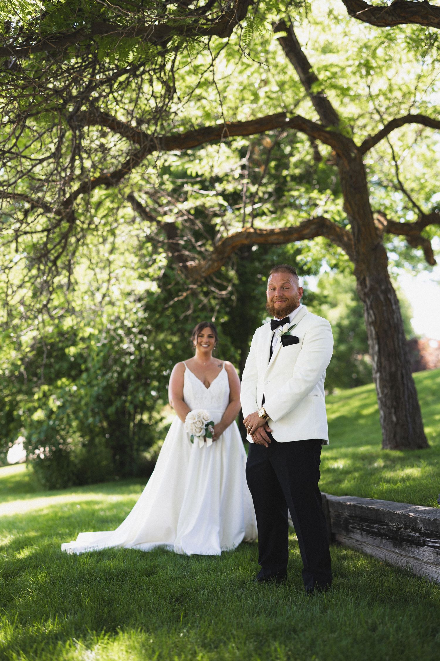 Newlyweds pose for a portrait under large trees in a garden setting.