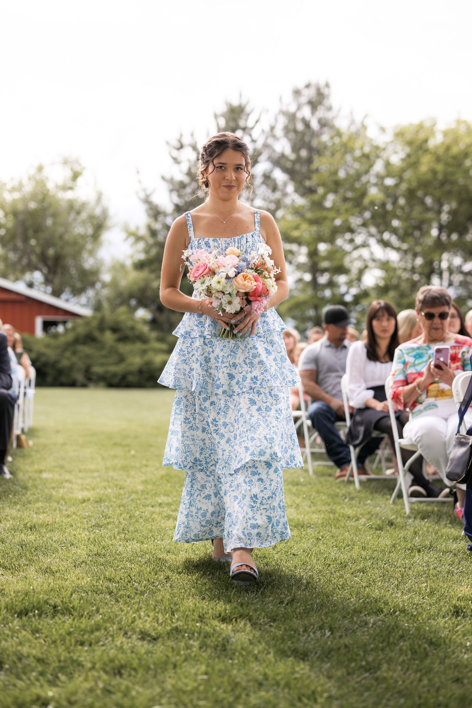 A person in a blue floral dress carries a colorful bouquet down the aisle at an outdoor wedding.