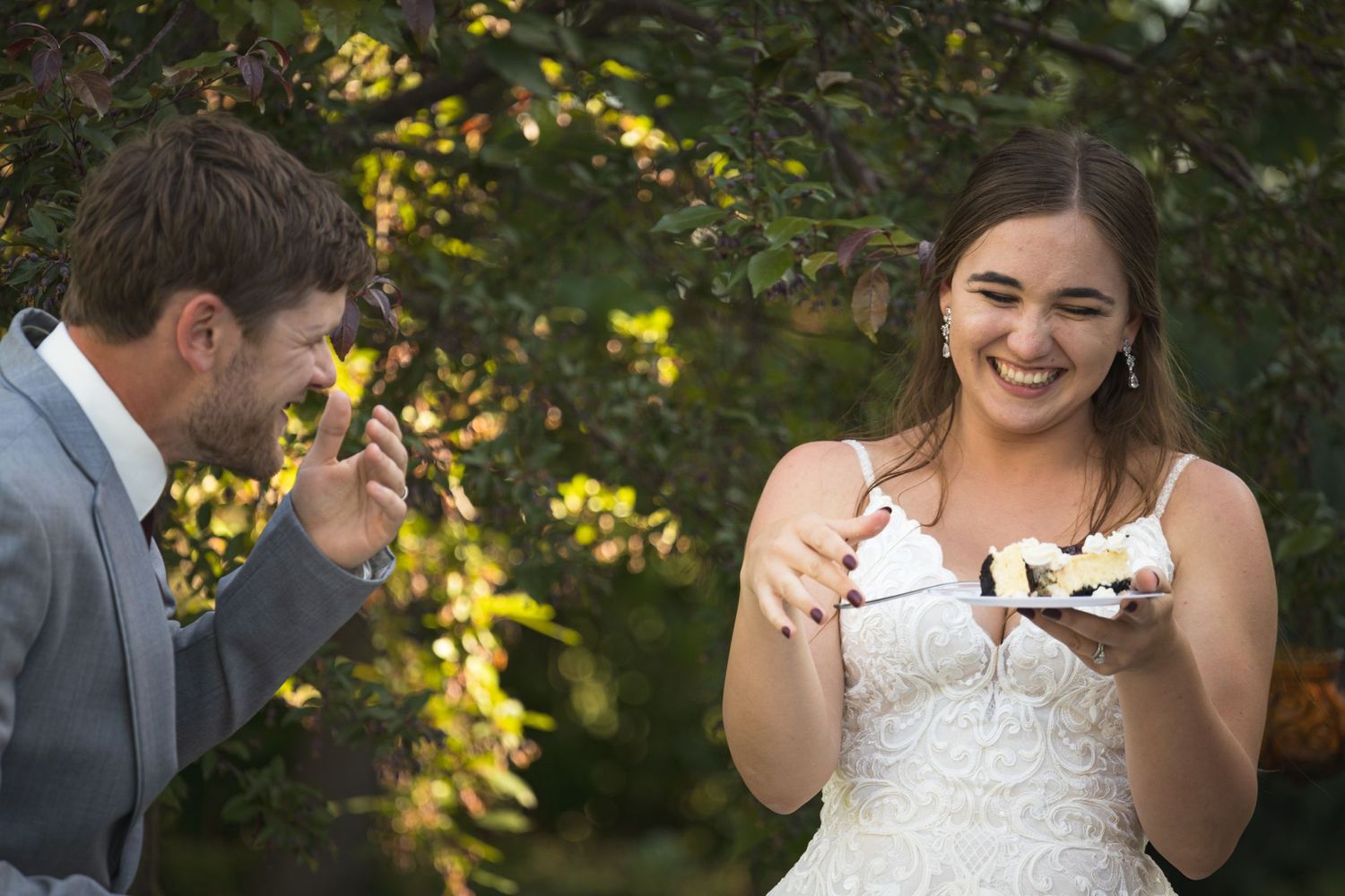 A couple laughs while sharing their wedding cake during an outdoor celebration.