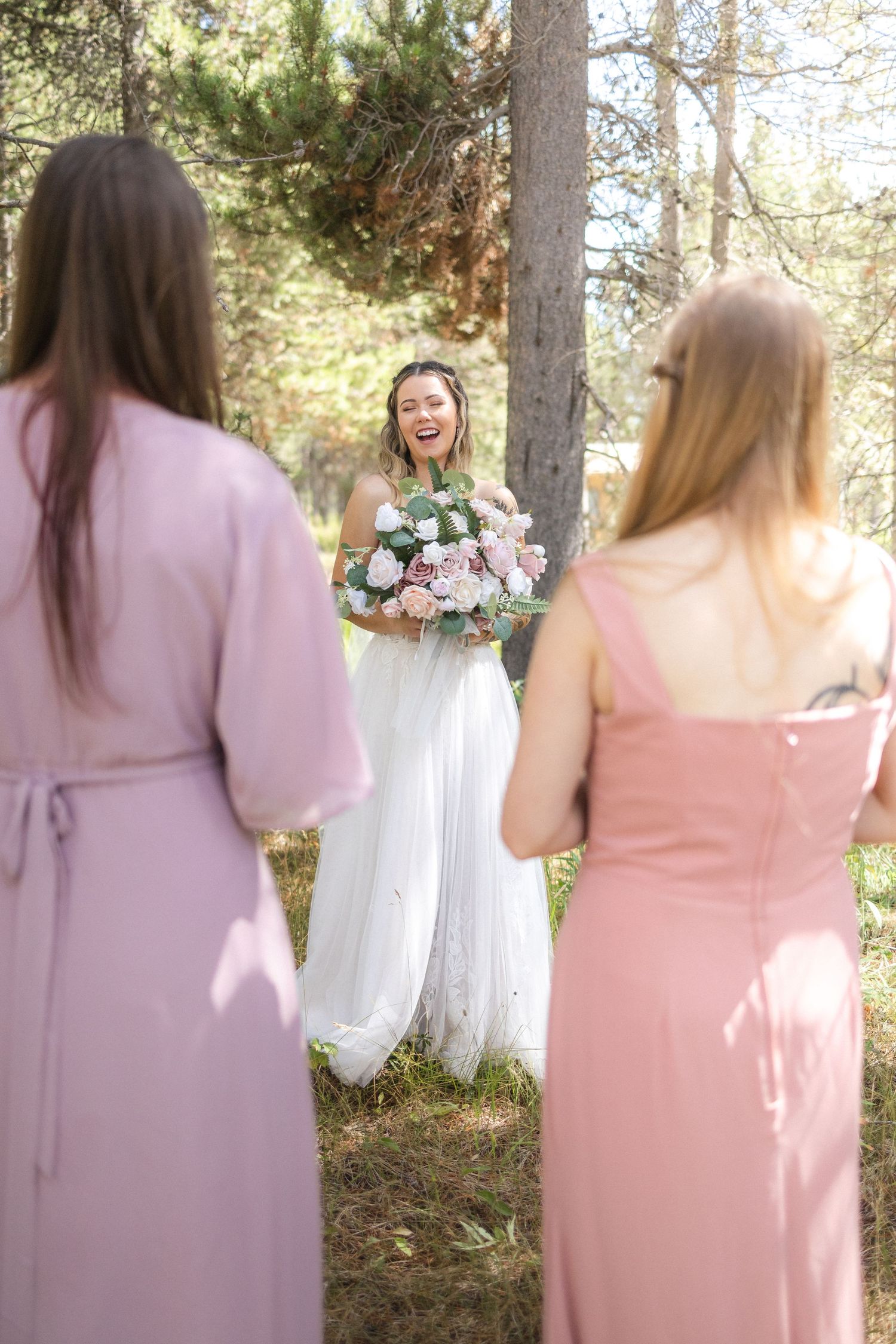 Attendants in pink dresses watch person in white wedding gown holding bouquet in forest.