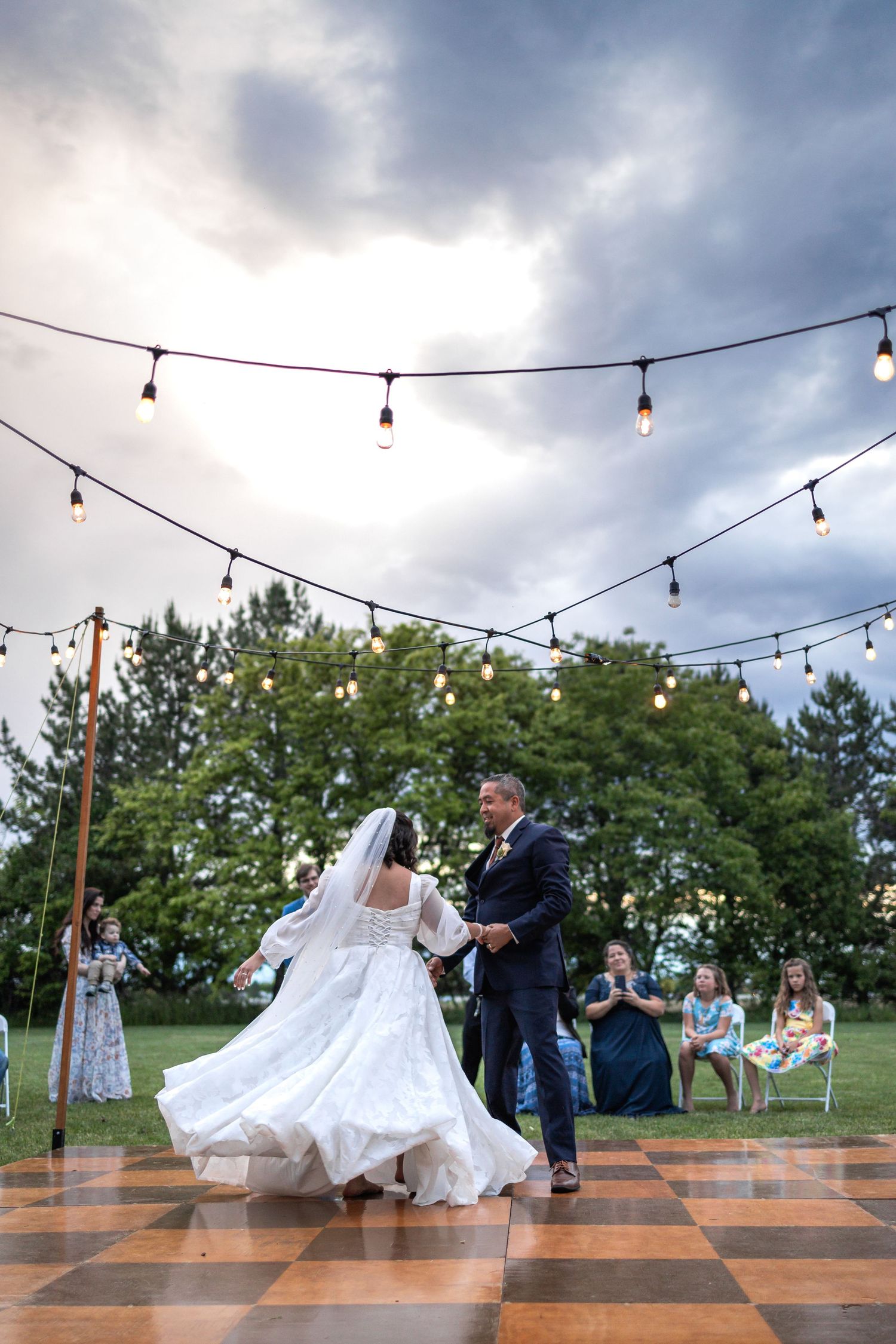 Newlyweds share a dance under twinkling lights on a checkered outdoor dance floor at dusk.
