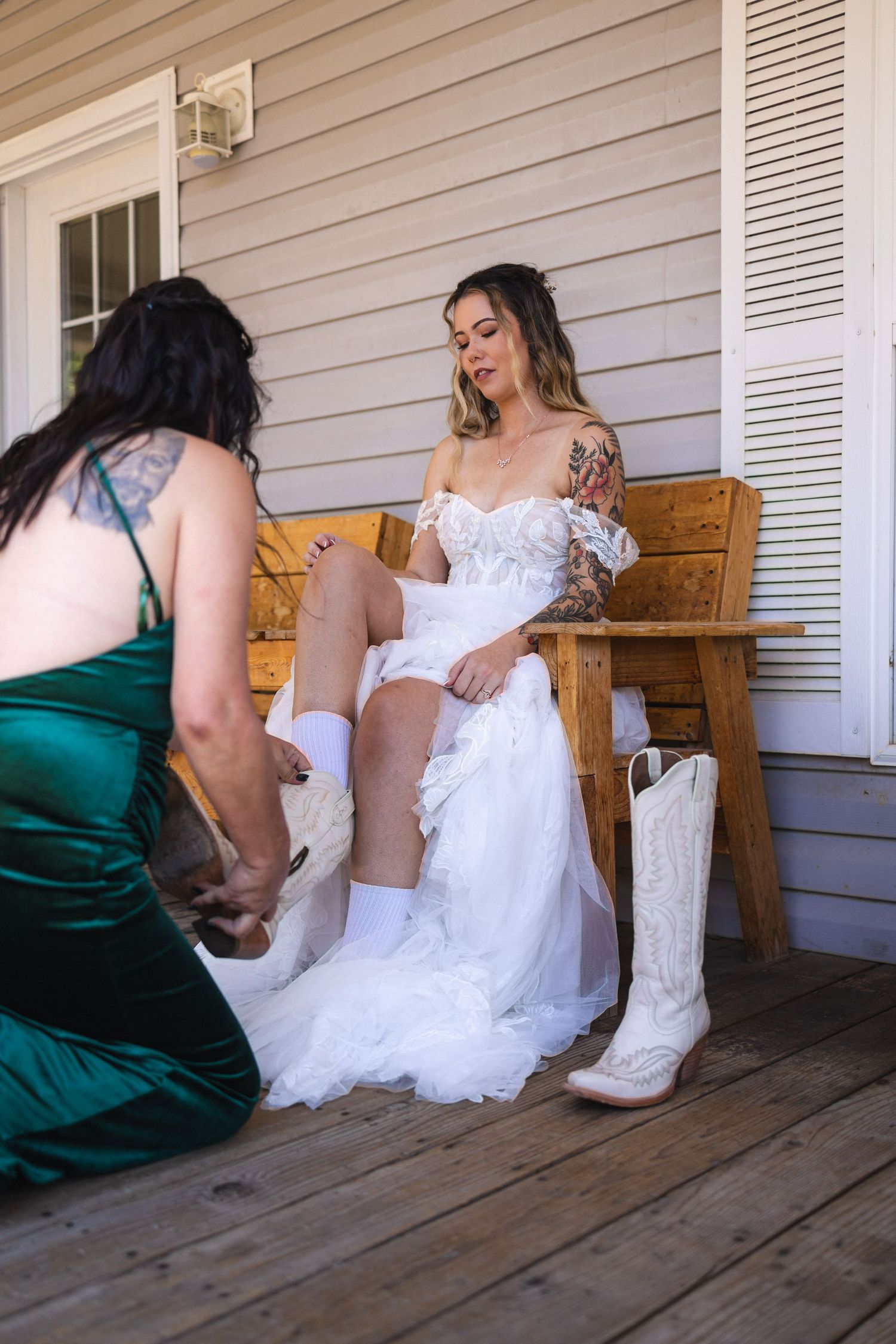 Helper in green dress helping bride put on white cowboy boots while sitting on wooden bench.