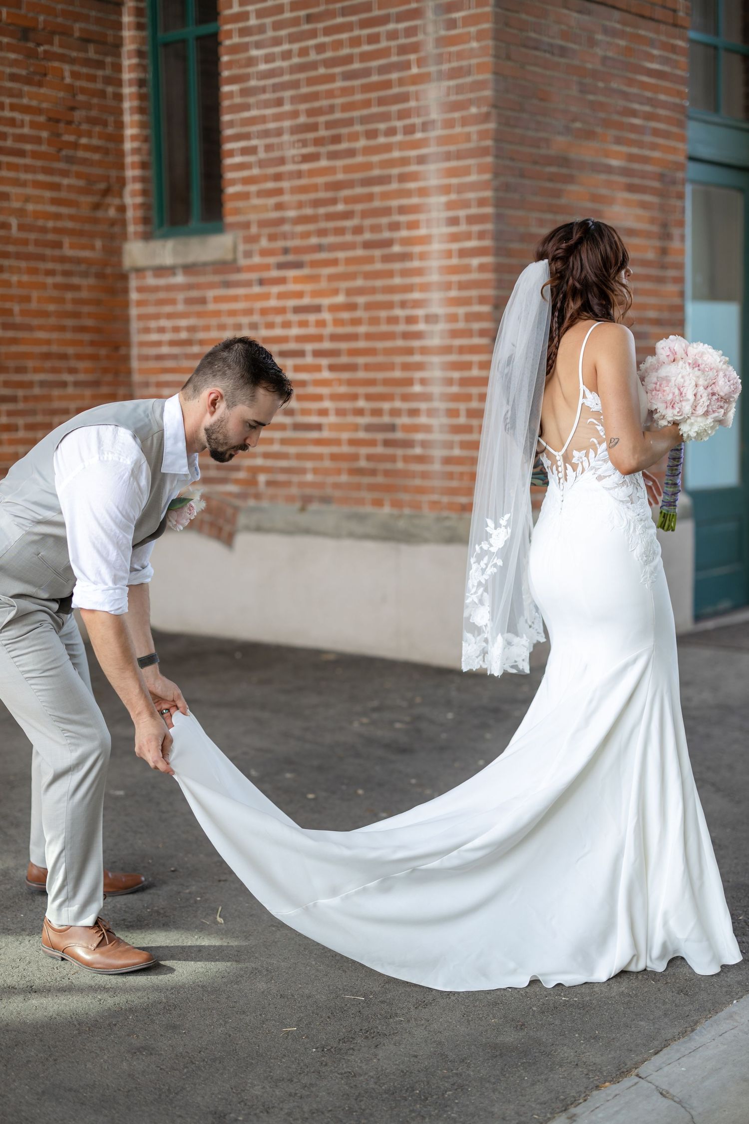 Groom helping with bride's train against rustic brick building exterior.