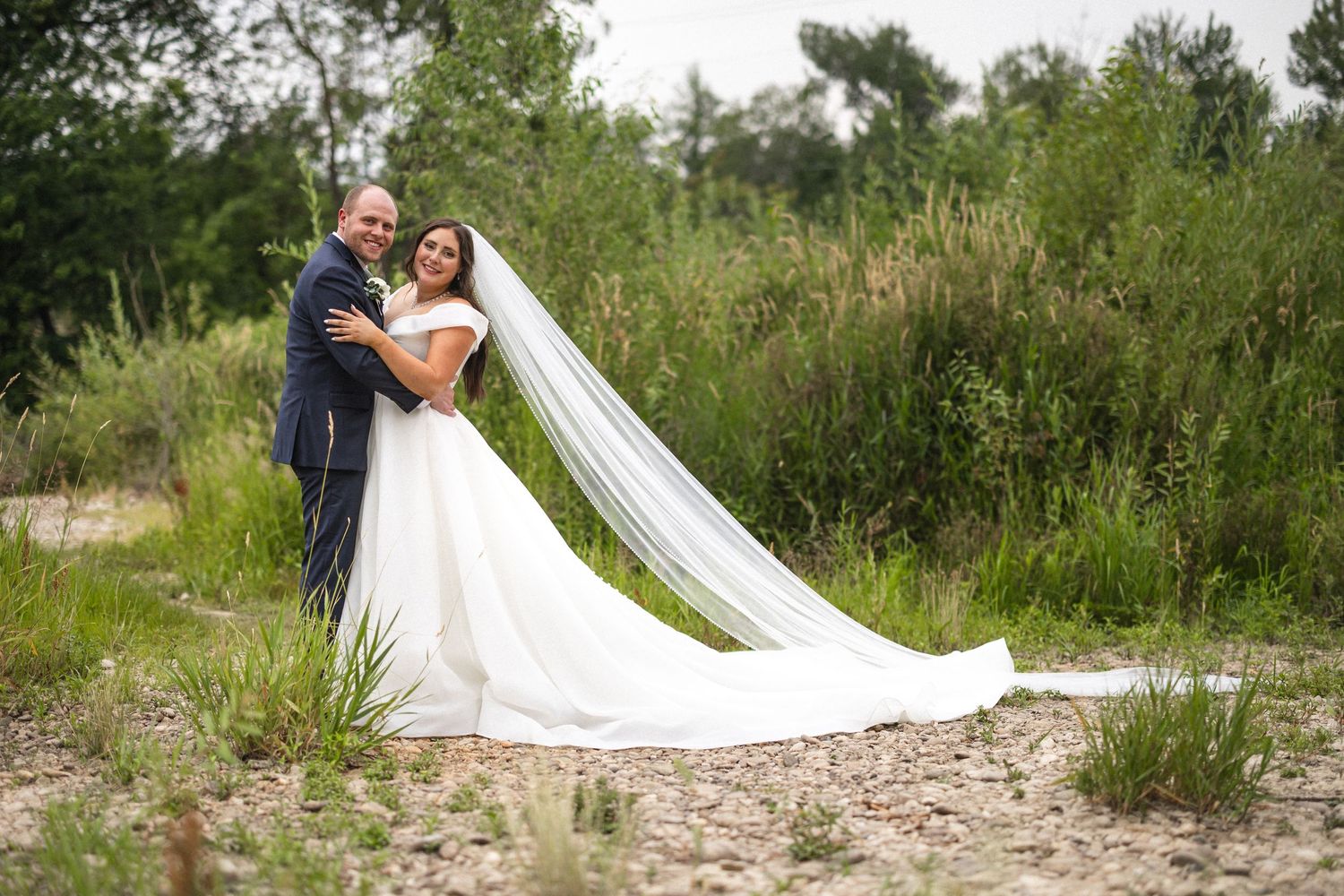 Wedding couple embraces on natural path with long flowing veil trailing behind.