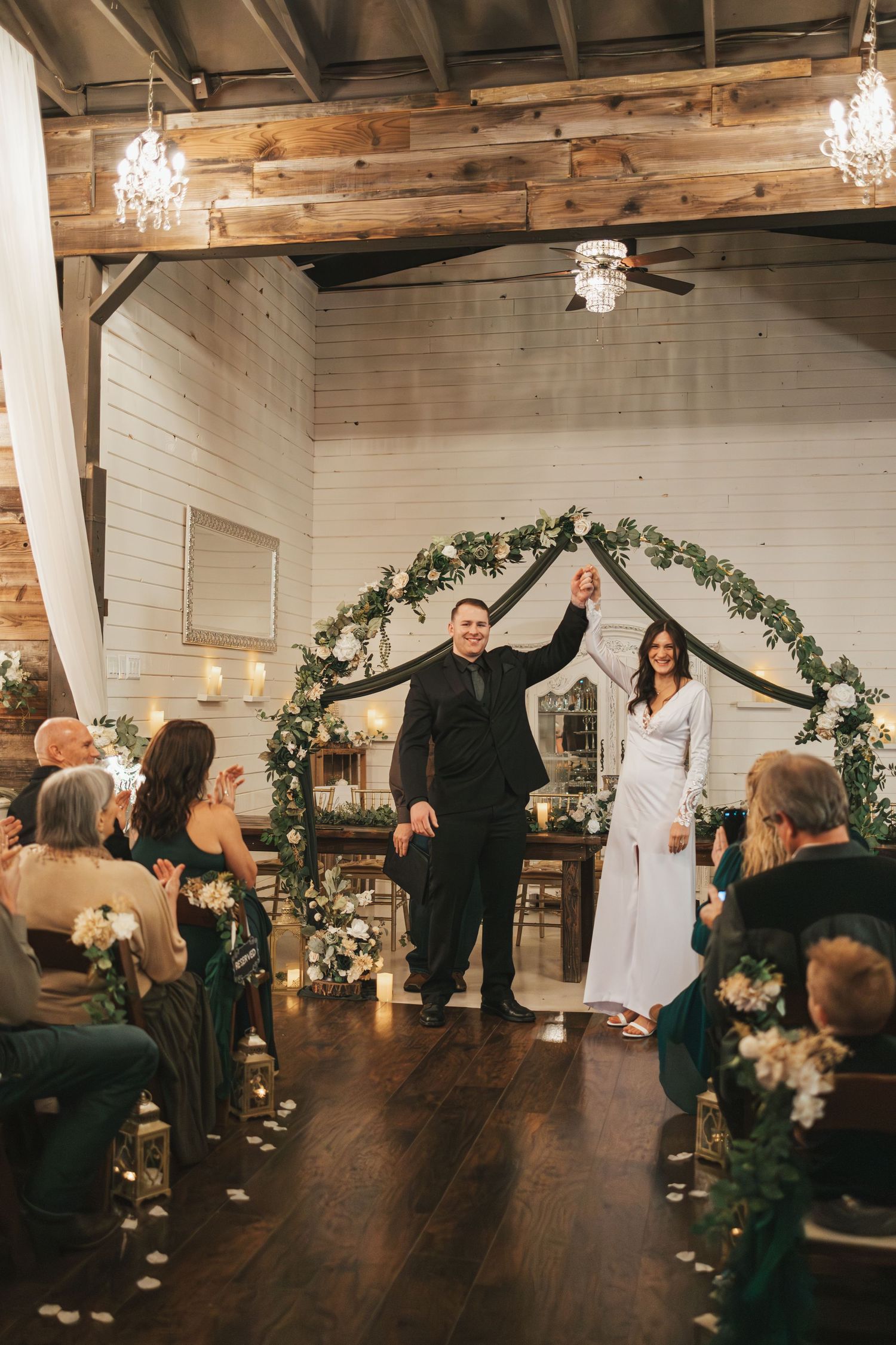 Newlyweds celebrating under floral arch in rustic venue with chandelier and wooden details.
