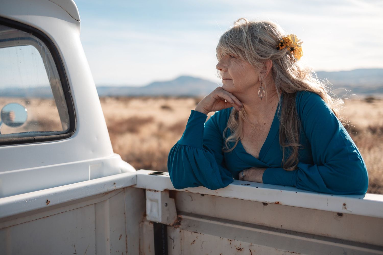 Person in teal shirt leans out of car window while driving through desert landscape at sunset.