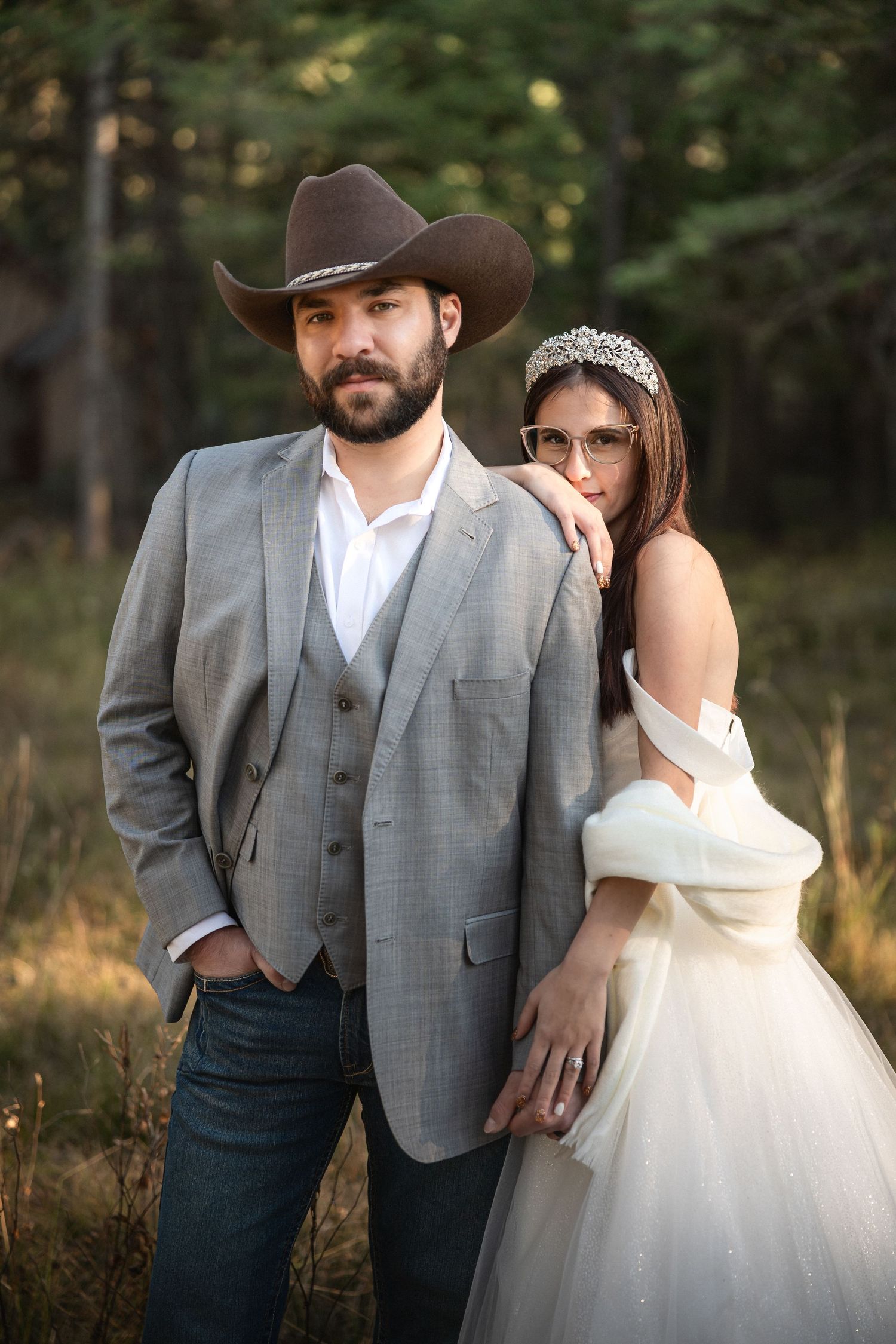 Bride in white gown and tiara poses with groom in gray suit and cowboy hat outdoors.