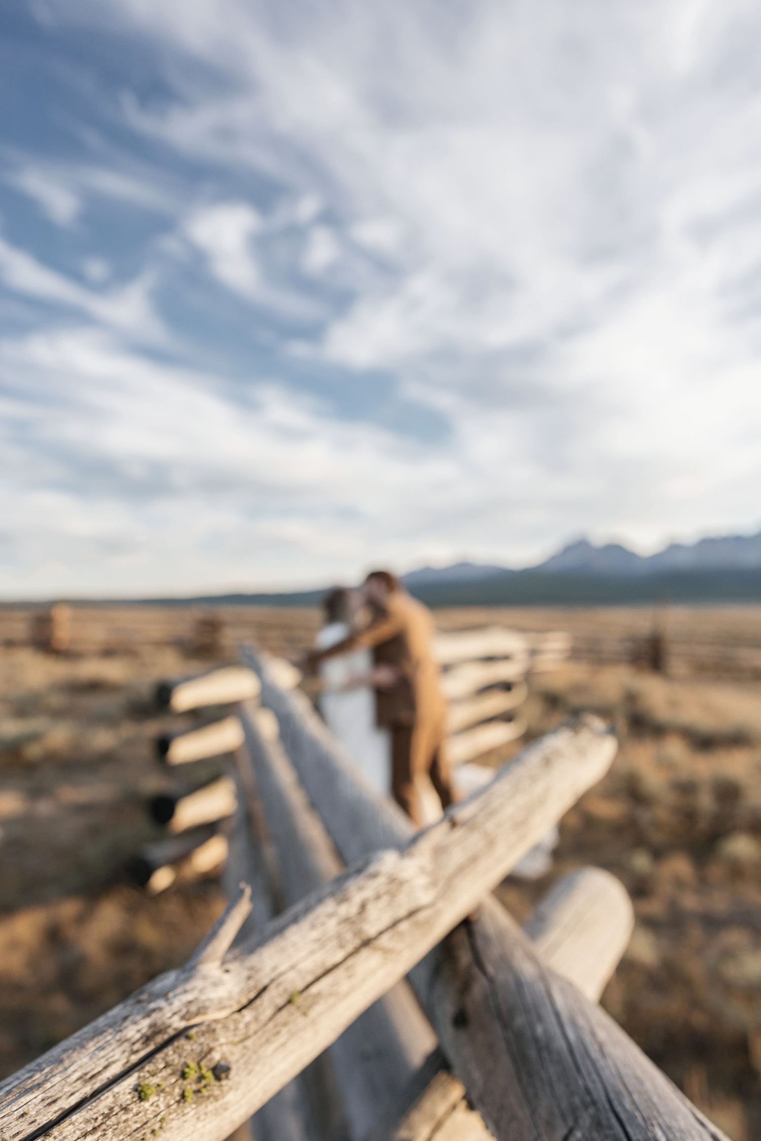 An artistic blurred photo of a couple by wooden fence rails with mountains in background.