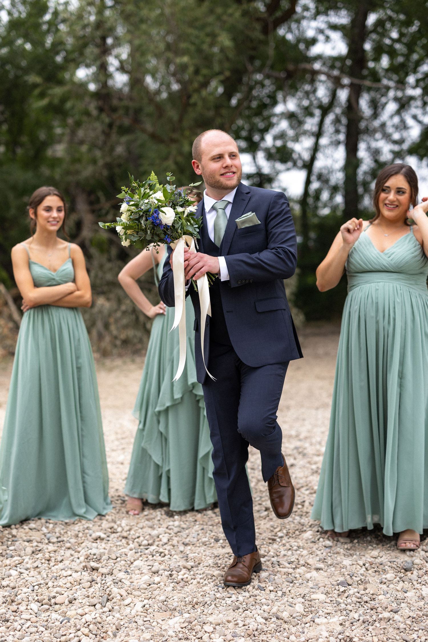 Groomsman carries bouquet while bridesmaids in sage dresses look on with amusement.