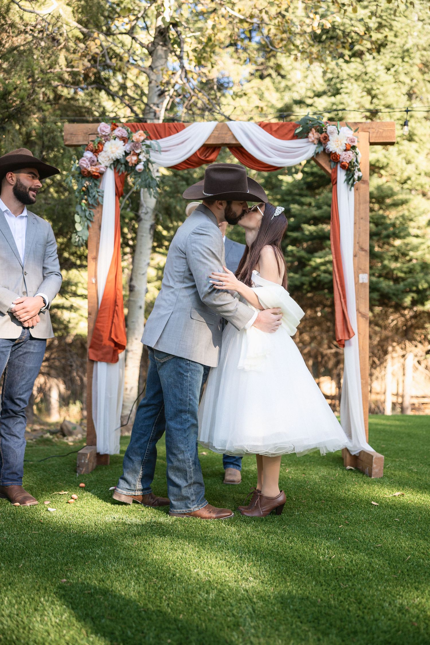 Newlyweds sharing first kiss under floral arch with wedding party looking on.