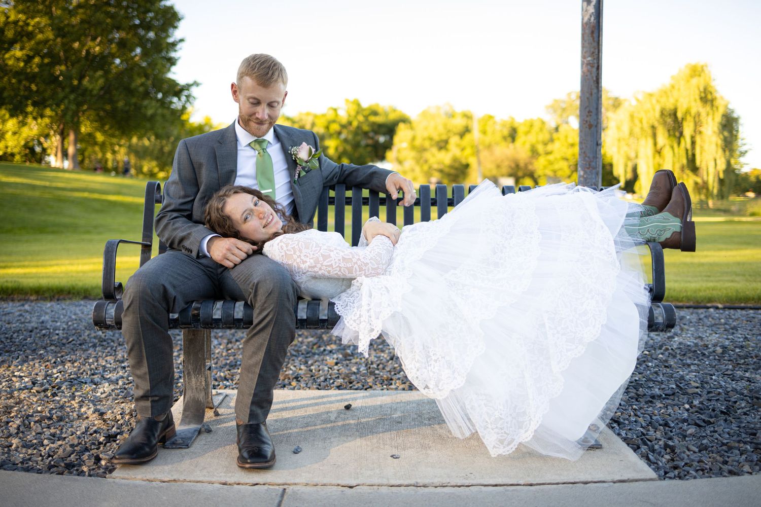 Playful wedding portrait of couple on park bench with bride's dress flowing.
