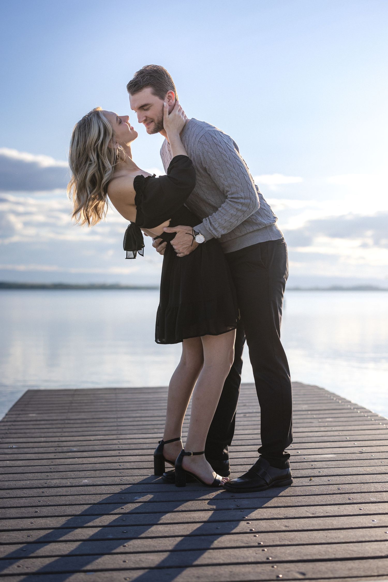 A romantic couple embraces on a wooden dock by a serene lake during sunset.