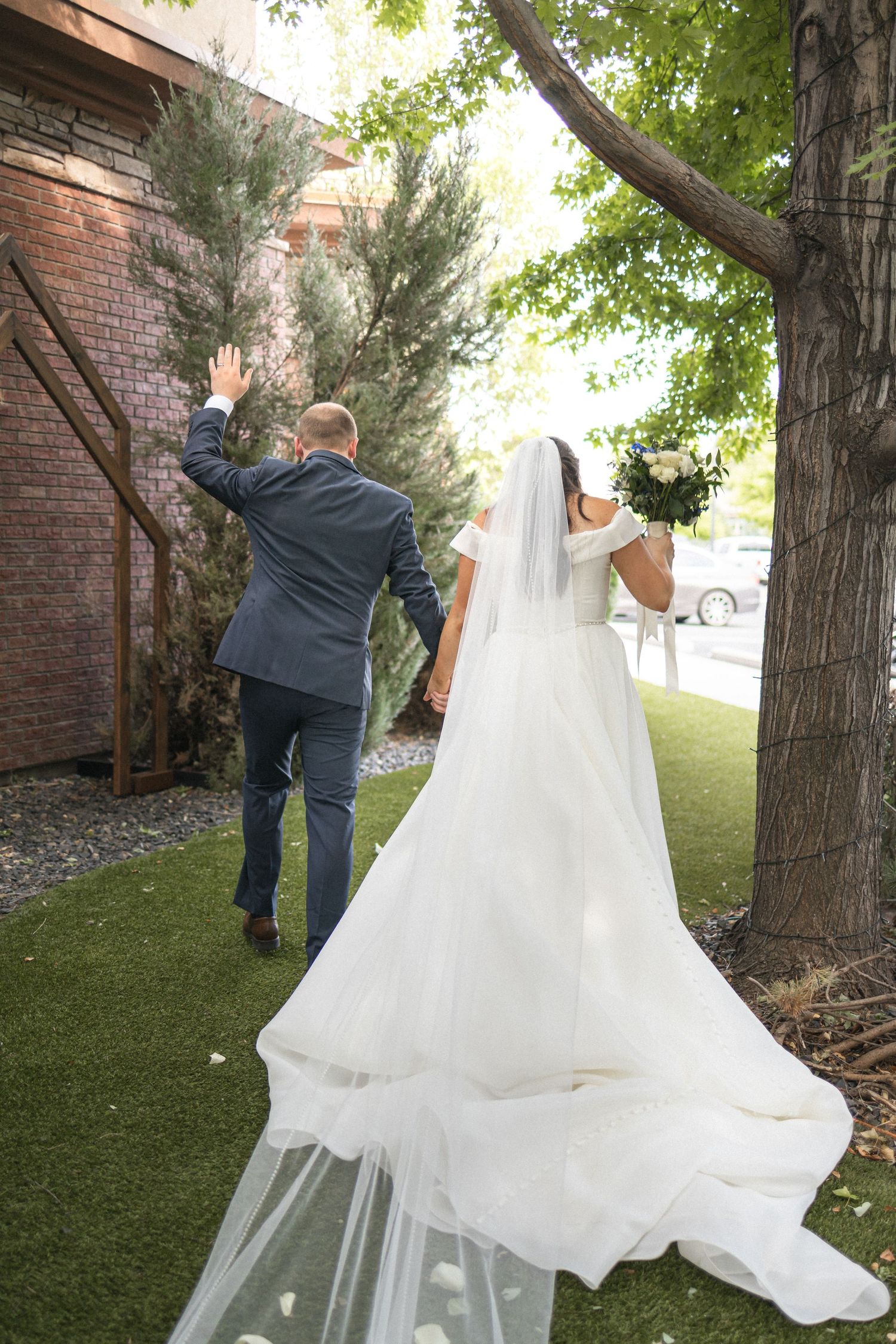 A wedding couple walking outside on artificial grass near a brick wall with the bride's flowing white dress trailing behind.