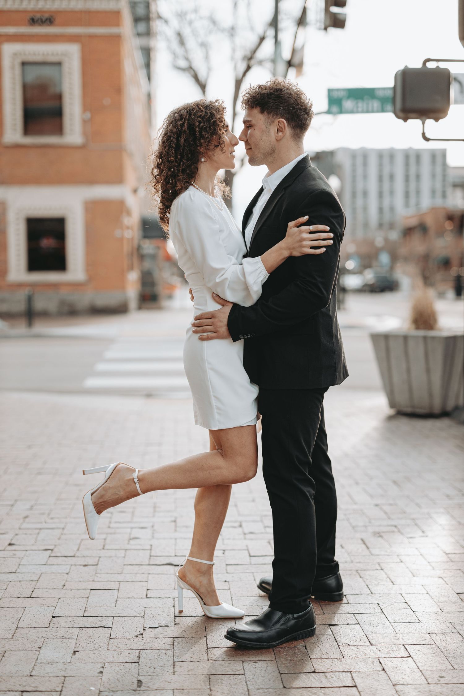 A couple in formal wear embracing while woman lifts leg in playful pose on city street.