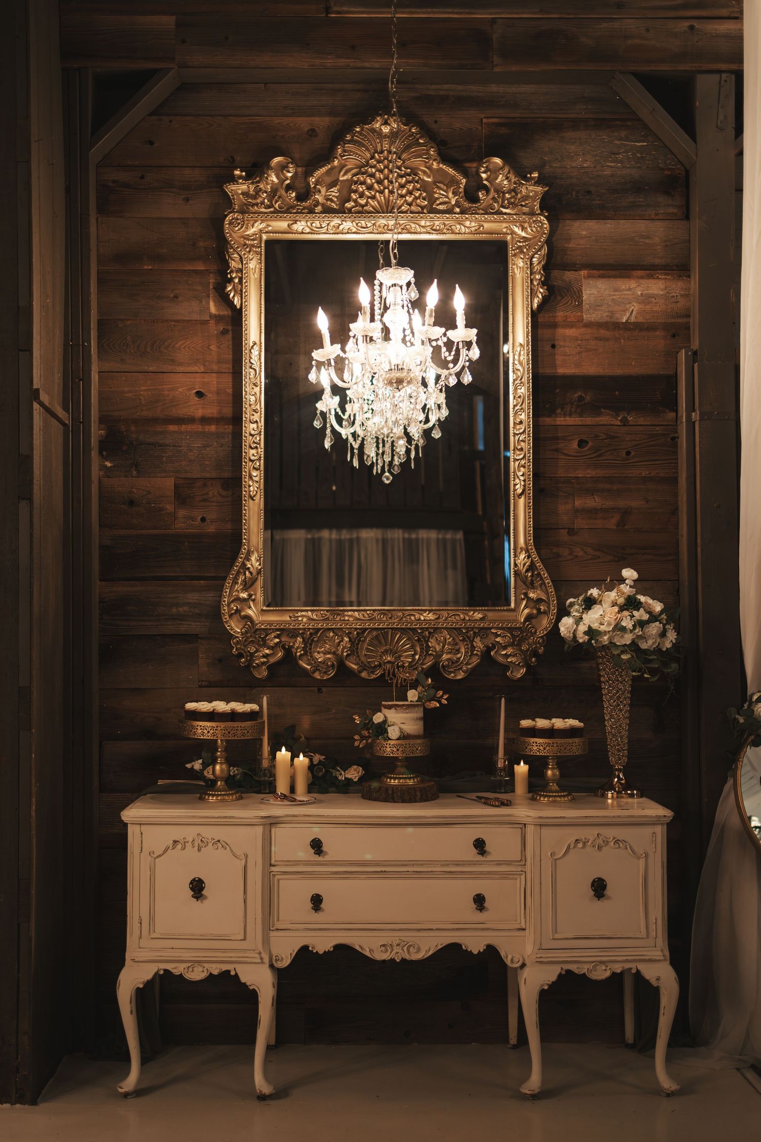 Elegant vintage white dresser with ornate gold mirror and crystal chandelier against rustic wooden wall.
