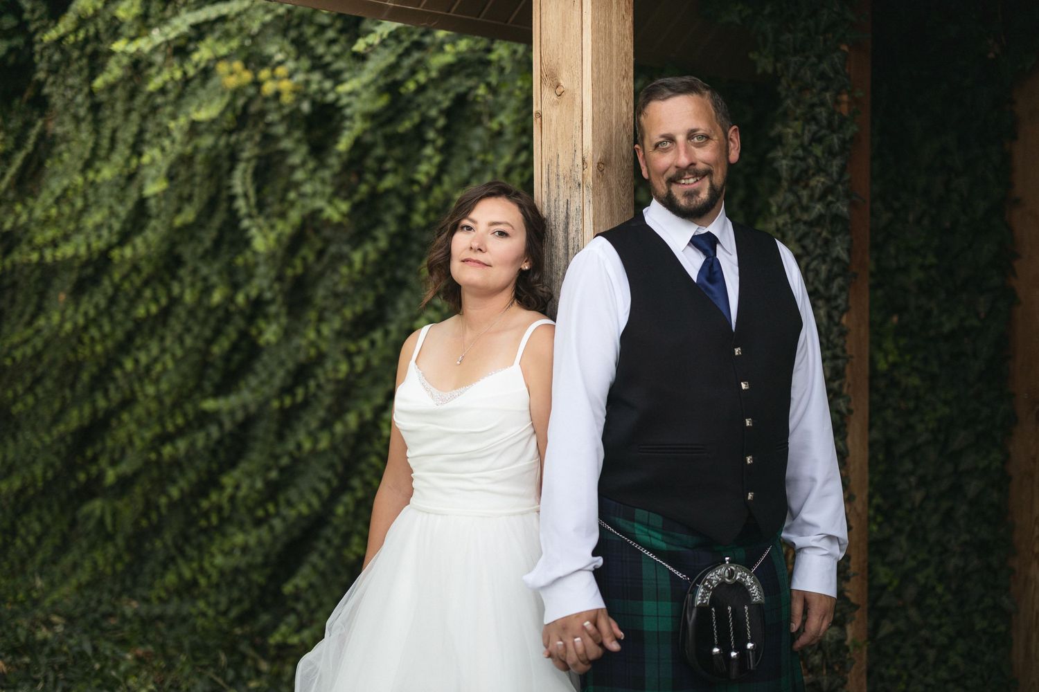 A couple in wedding attire pose together against a backdrop of lush green vines.