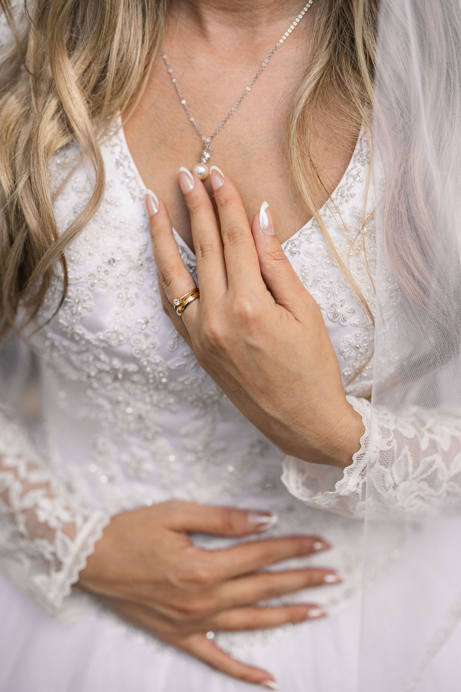 Bride's hands with wedding ring and necklace against white lace dress.