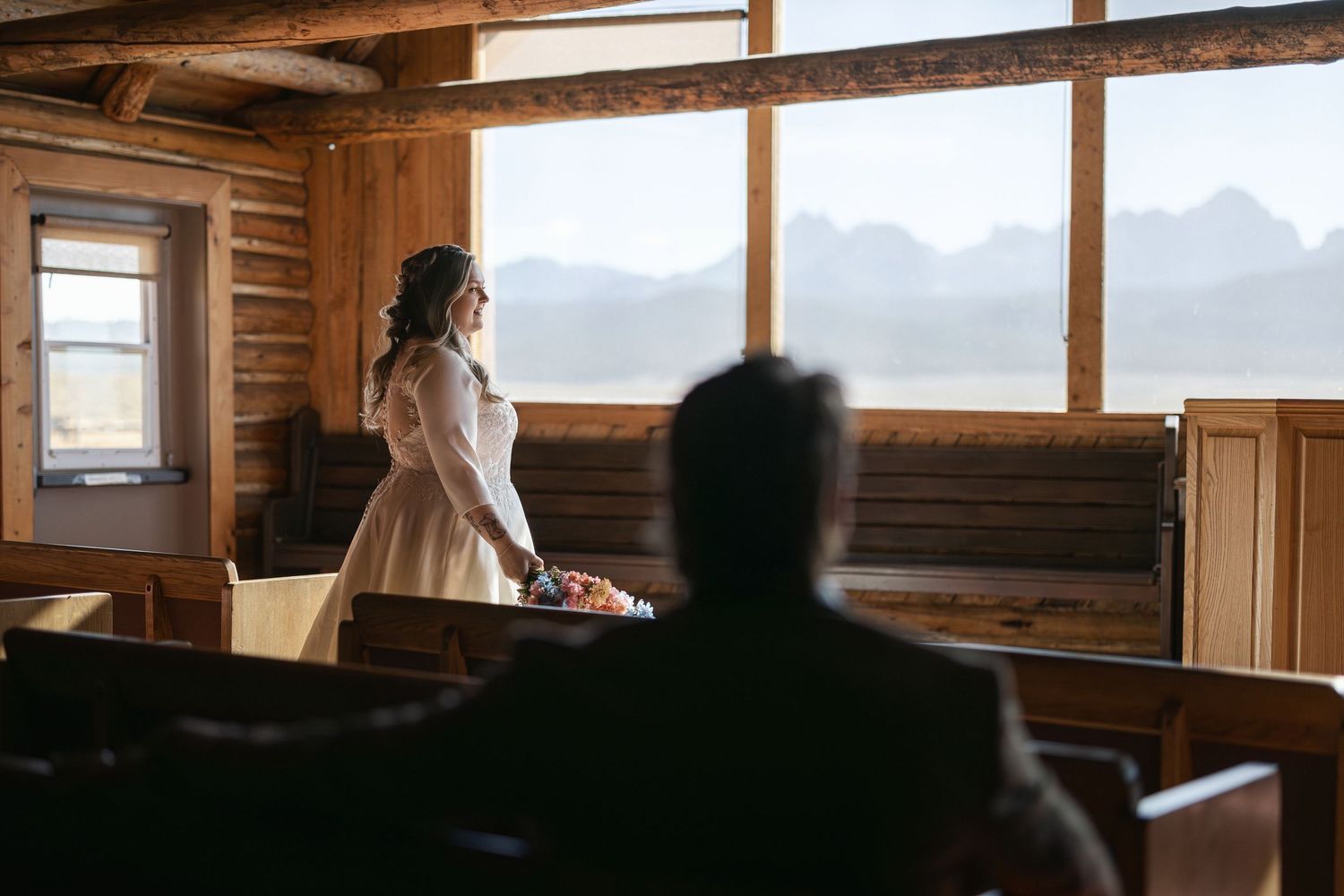 Sunlit chapel interior with wooden beams and mountain views through large windows.