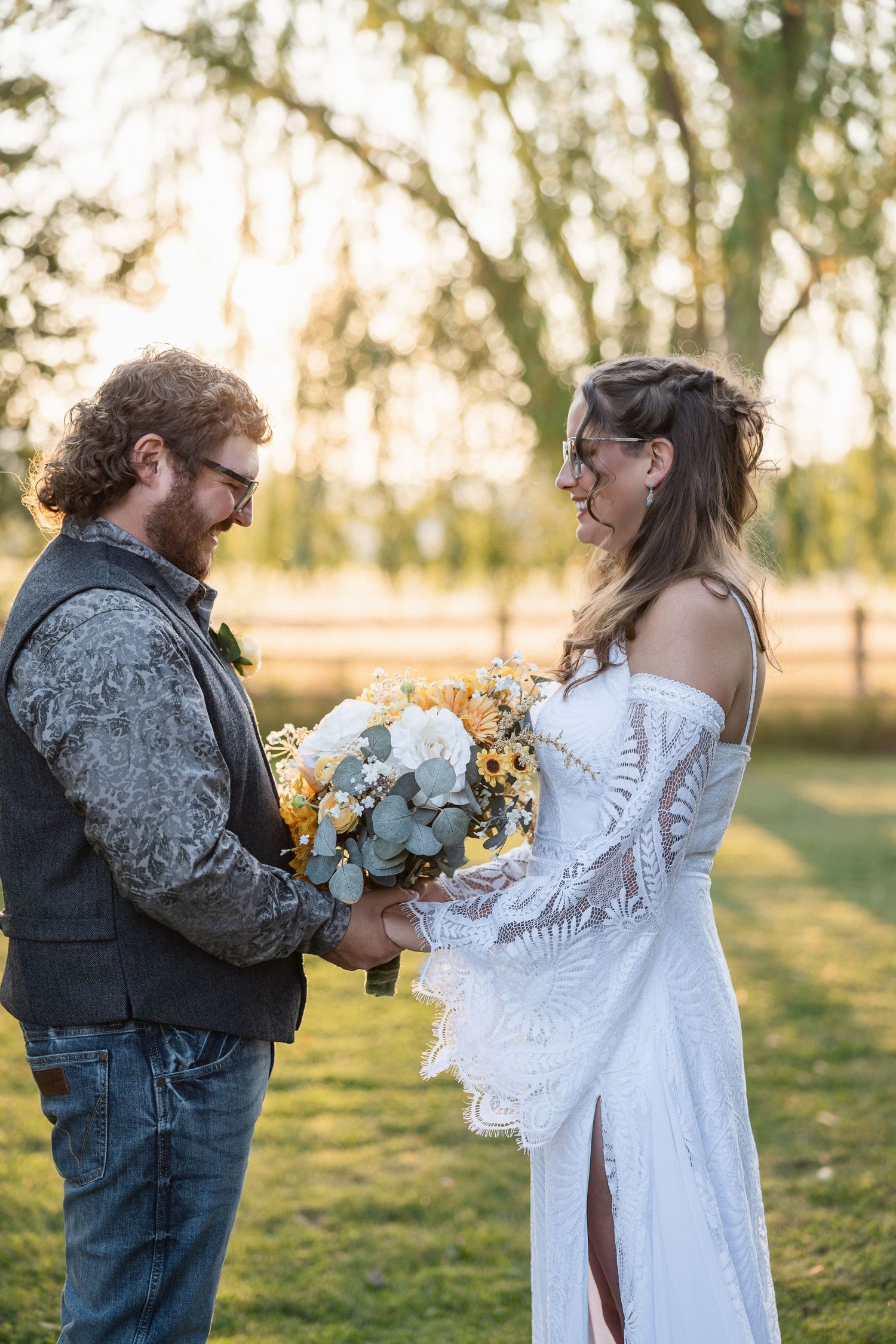 Couple in wedding attire sharing an intimate moment outdoors under willow trees at golden hour.