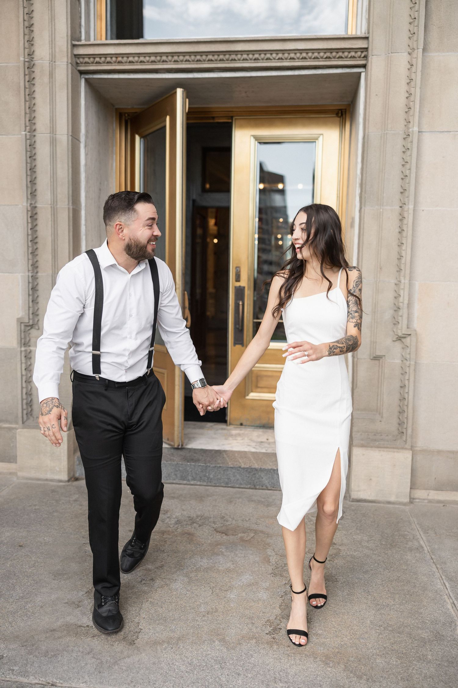 A stylish couple walks hand in hand outside a grand building entrance.