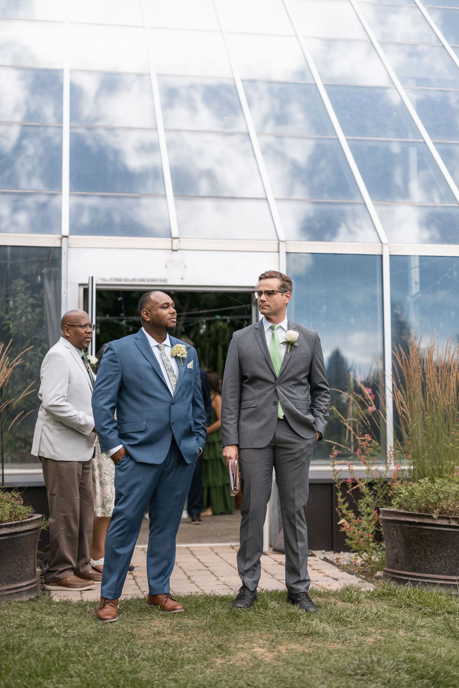 Groomsmen in suits standing outside greenhouse wedding venue.