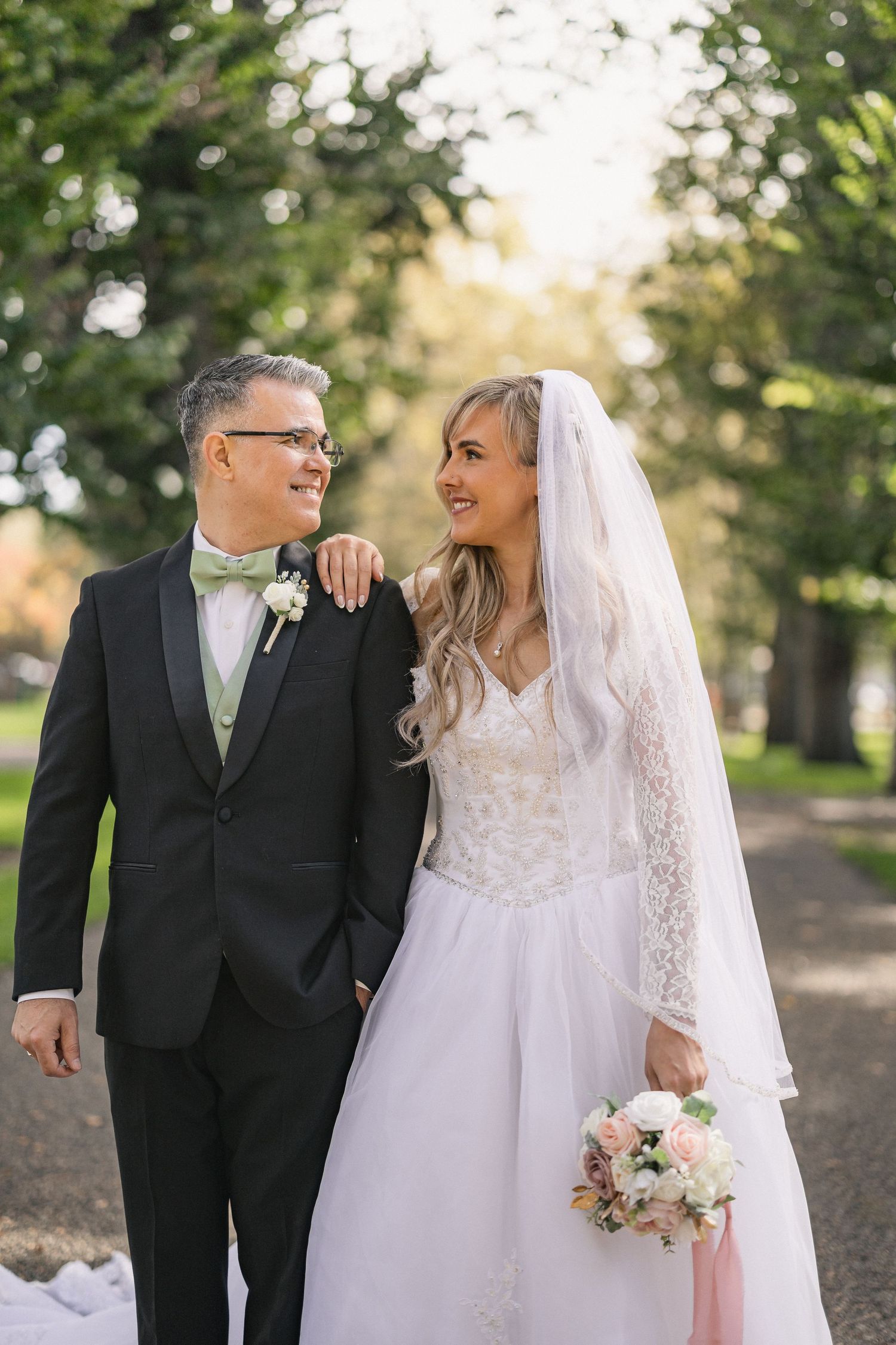 Happy newlyweds walking together outdoors in formal attire with veil flowing.