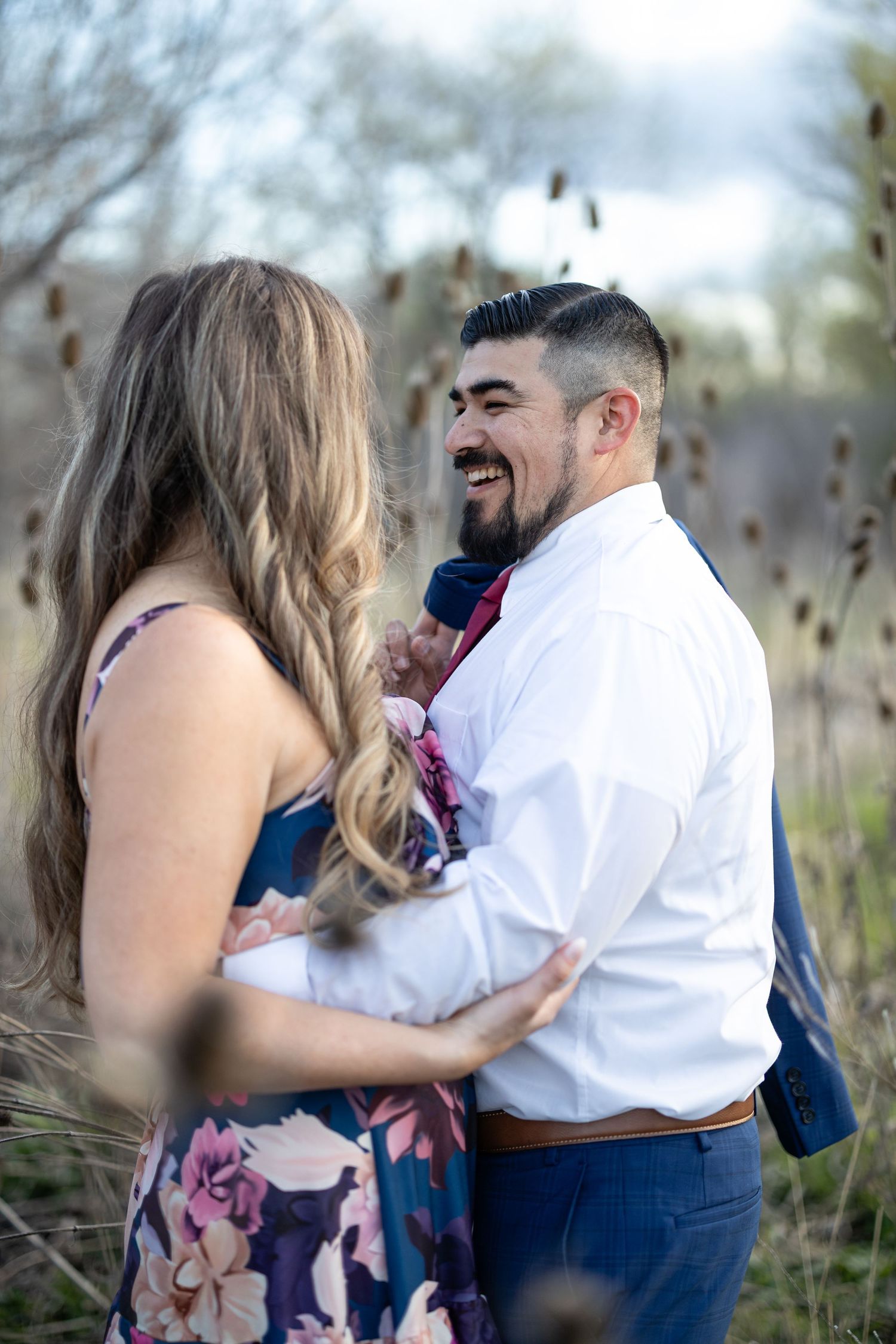 A romantic couple laughs together in a field of dried cattails during an outdoor photo session.