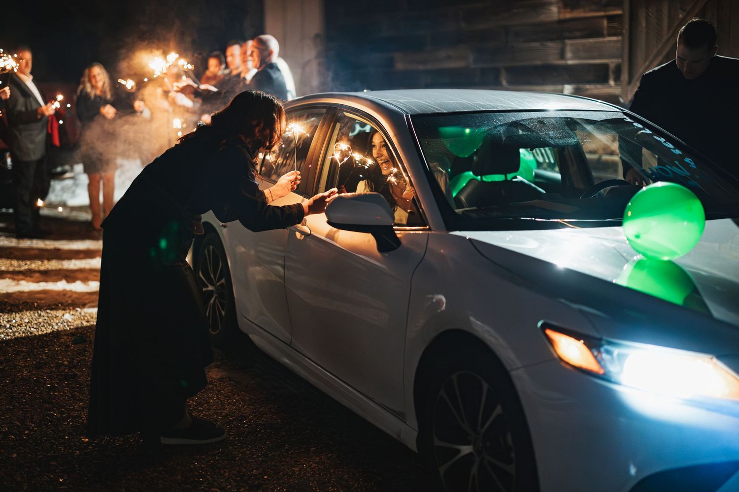 White car illuminated by sparklers during nighttime wedding departure celebration.