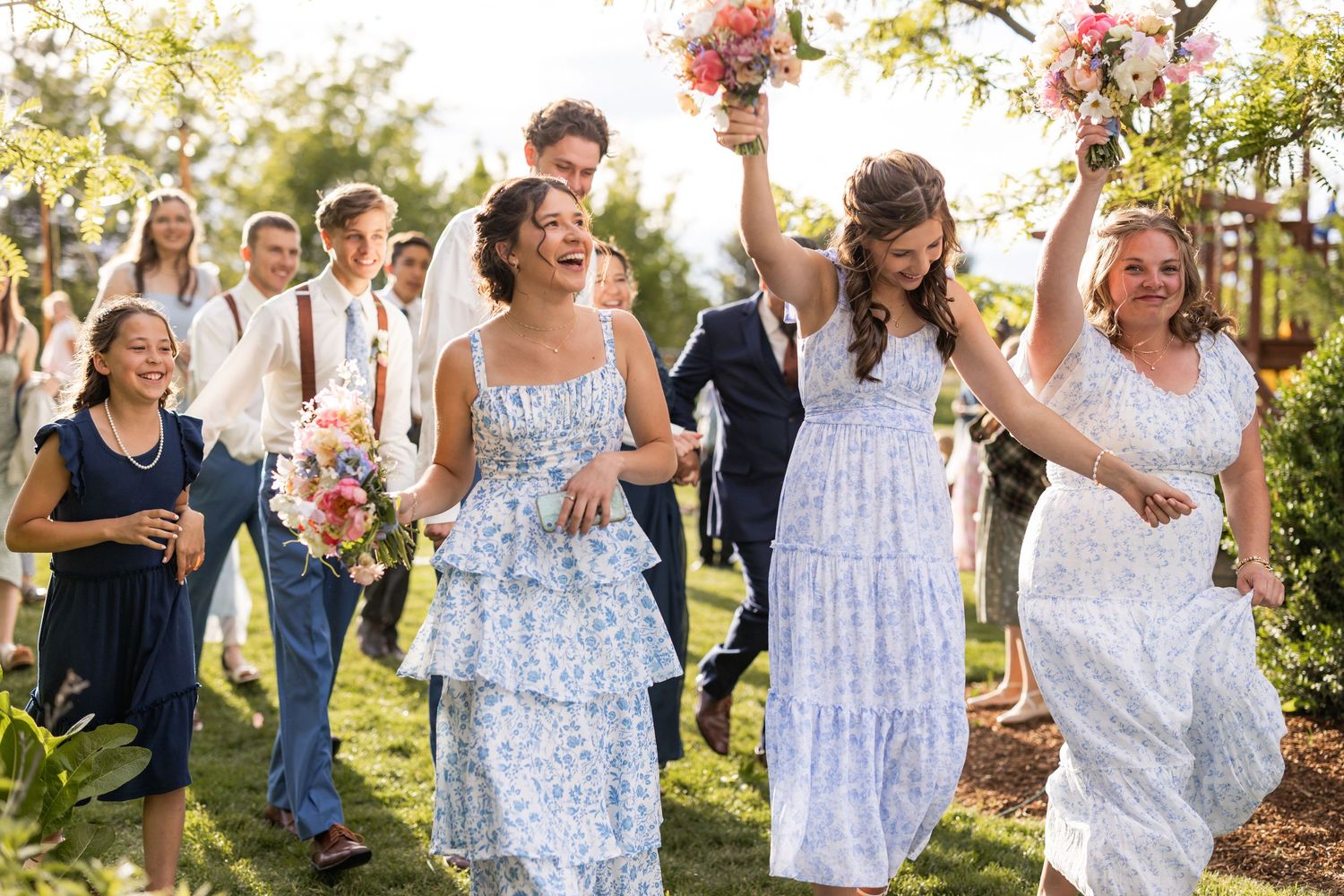 Wedding party in white dresses celebrates joyfully outdoors while holding floral bouquets.