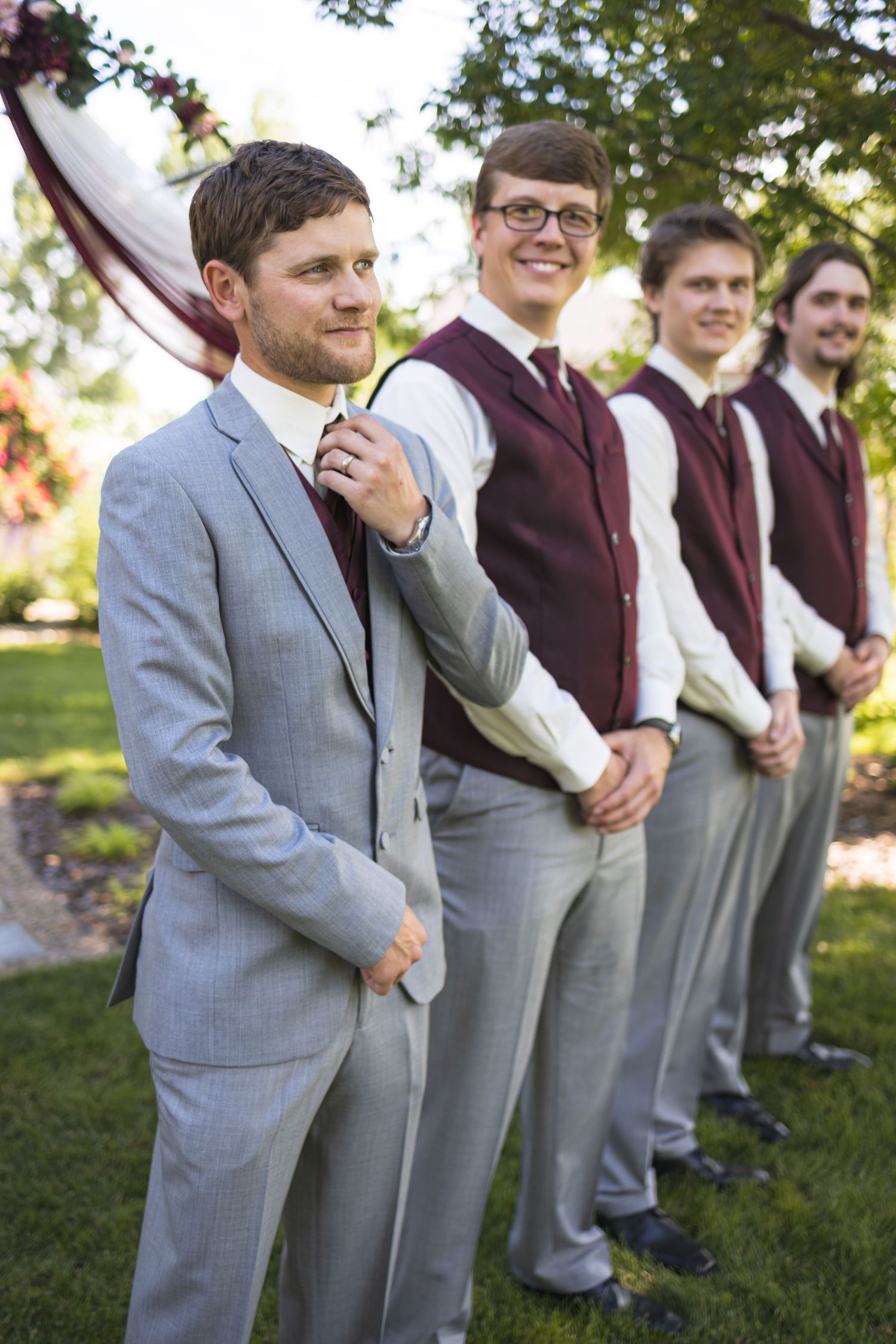 Groomsmen lined up in grey suits and burgundy vests at outdoor wedding.