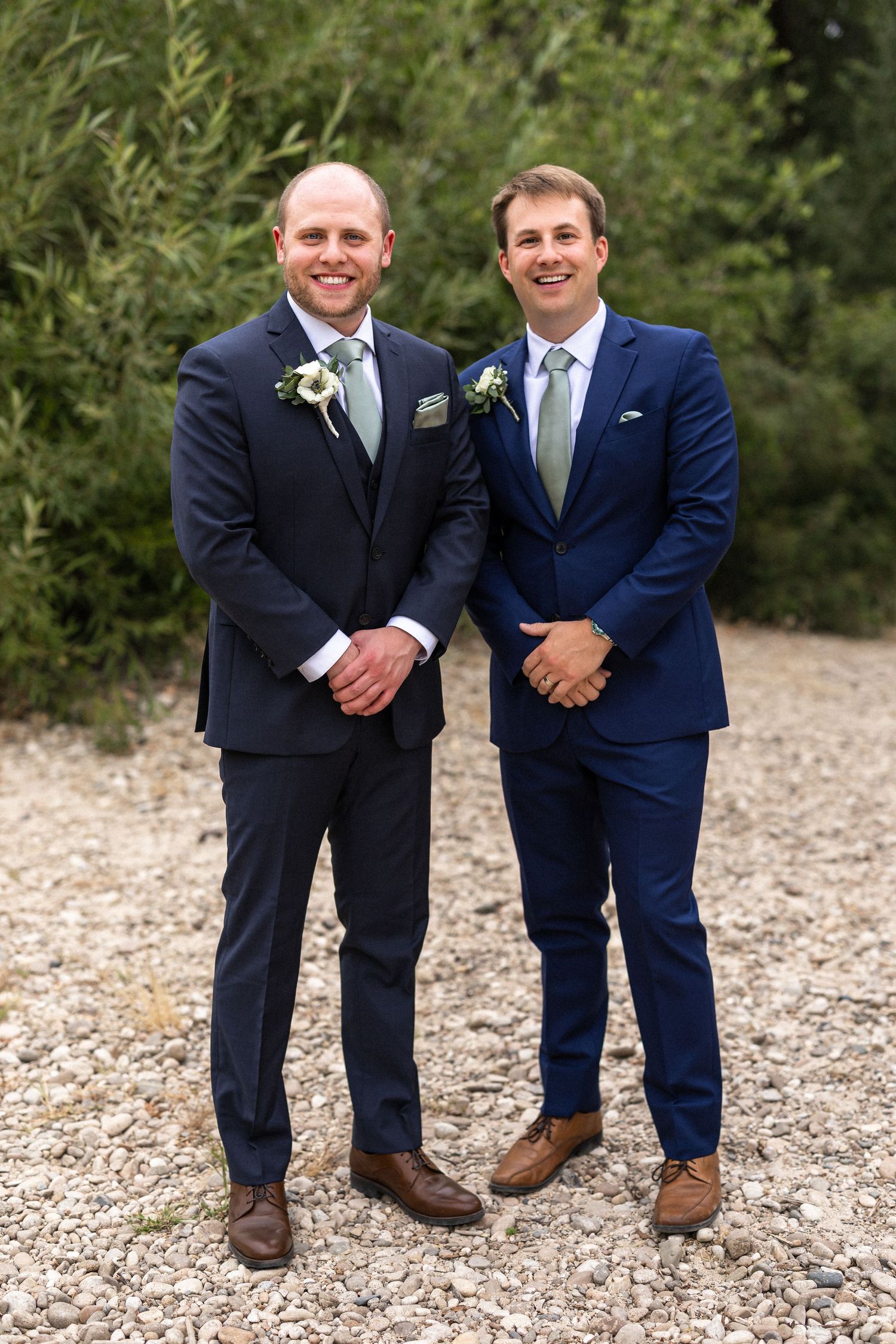 Two groomsmen in navy blue suits and brown shoes stand together on a gravel path with greenery in background.