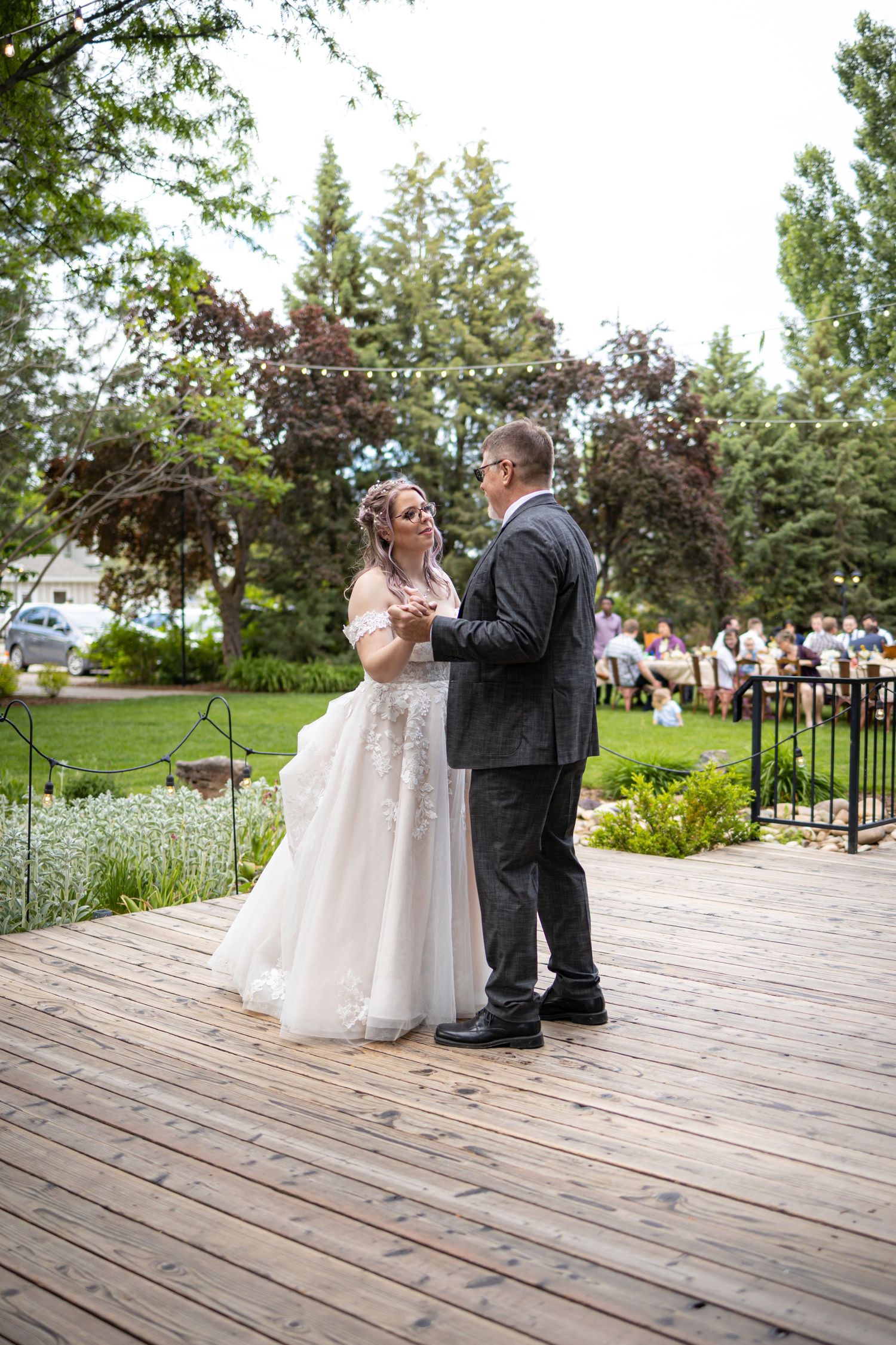 Couple dancing their first dance on a rustic wooden deck with guests watching in background.