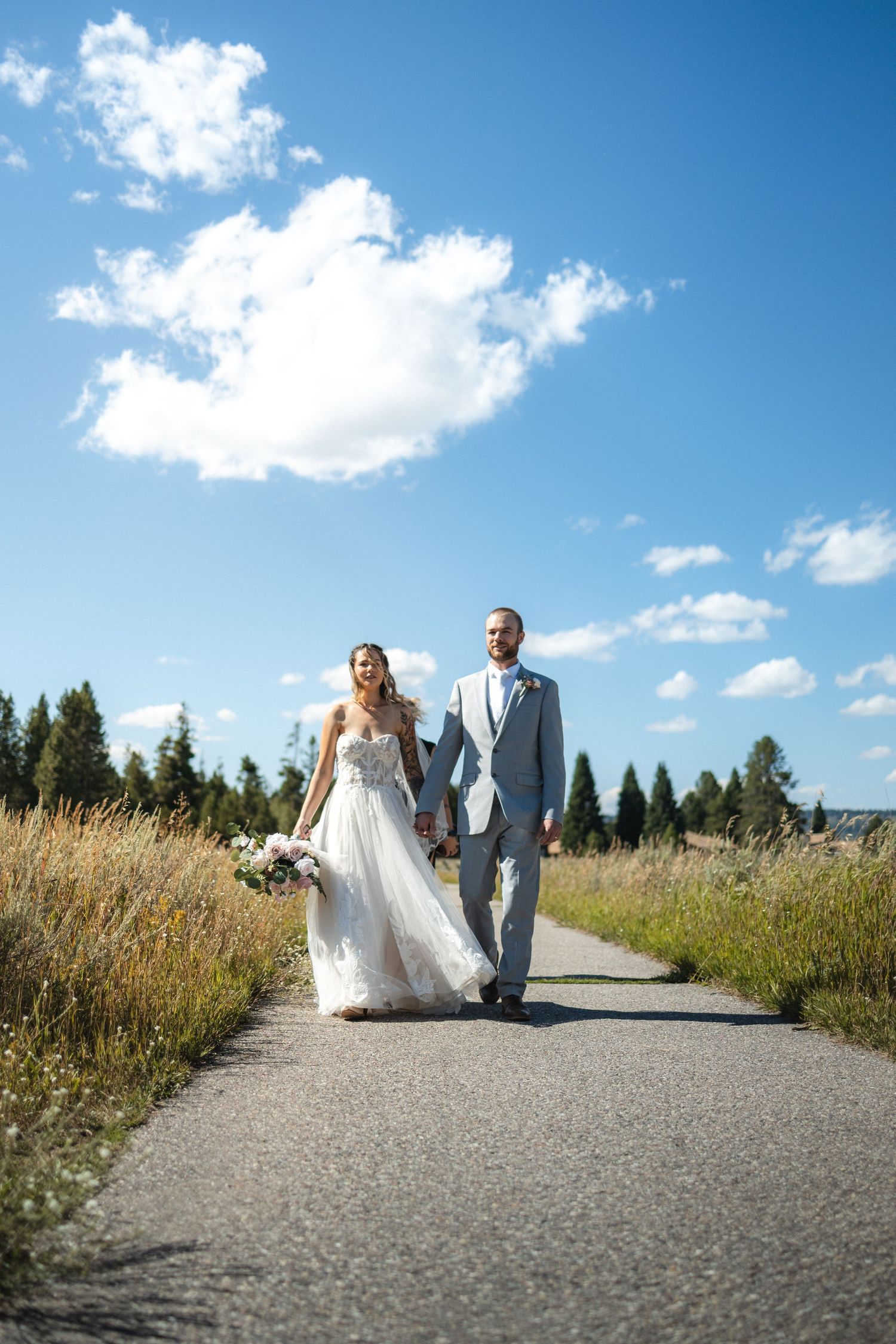 A couple in wedding attire walks along a path surrounded by tall grass and evergreen trees on a sunny day.