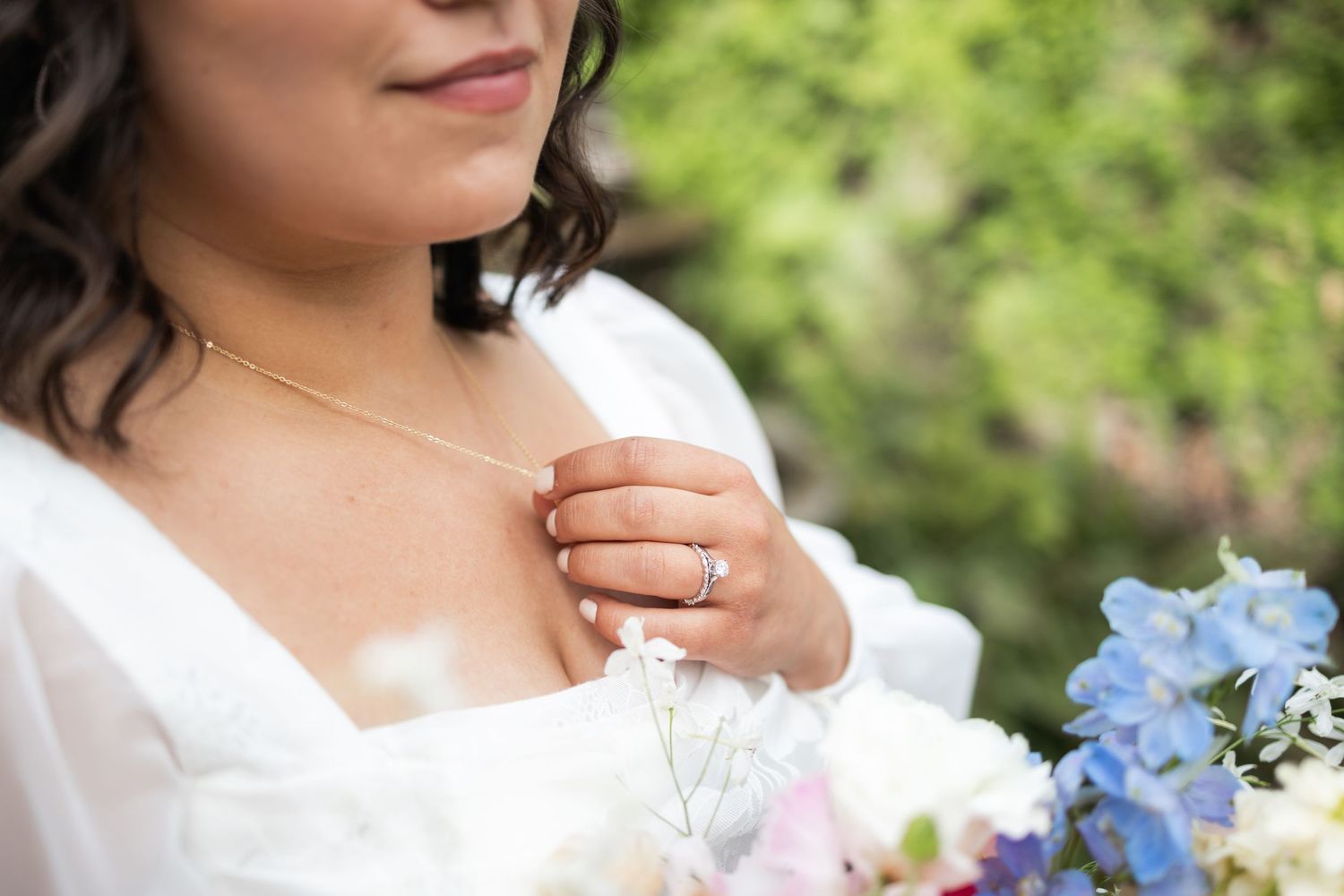 Close-up detail of wedding jewelry and white dress with blue flowers in bouquet.