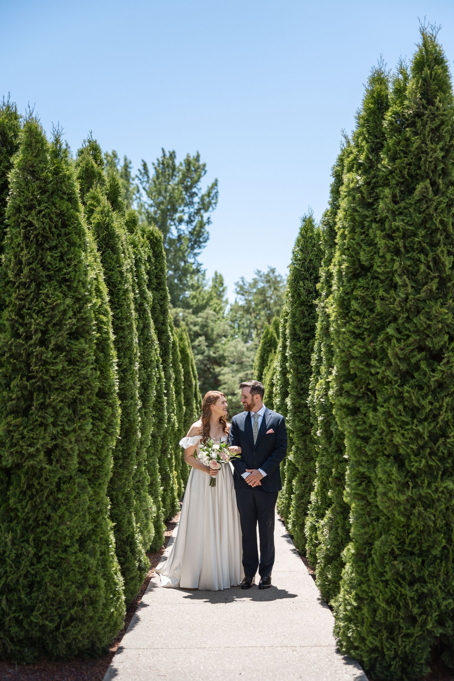A couple stands between tall cypress trees on a paved pathway in an elegant garden setting.