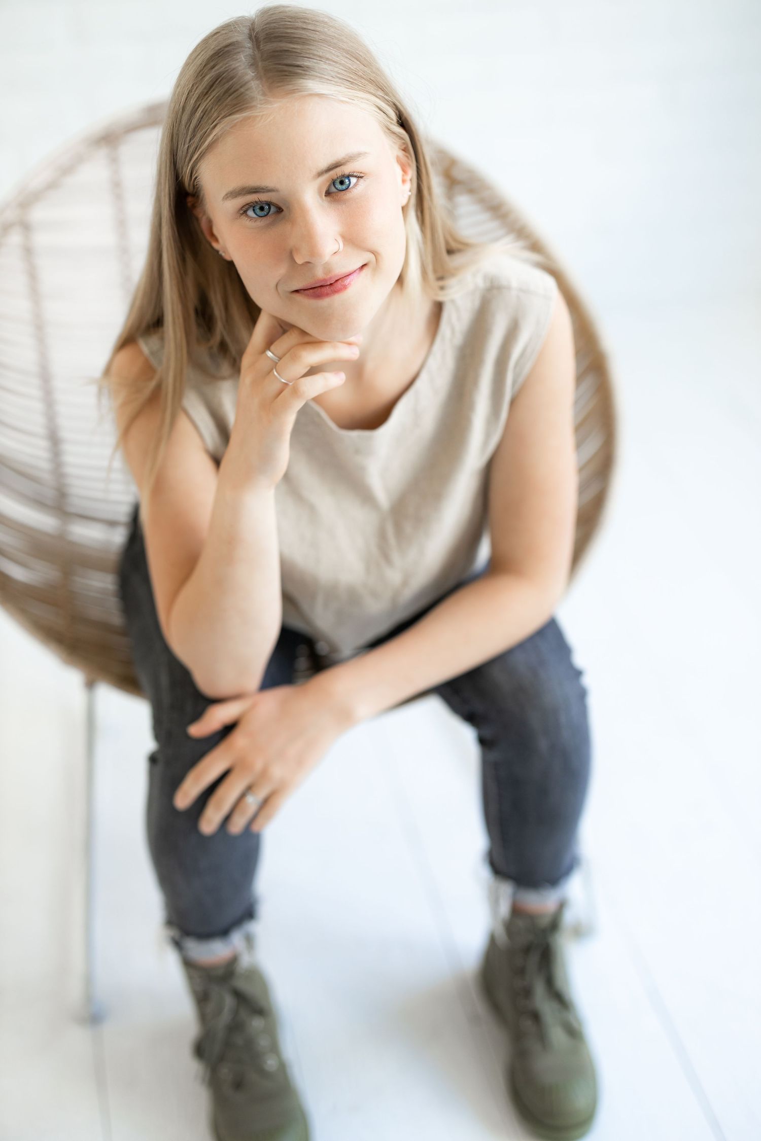 Person in casual attire sits on white floor wearing olive boots while resting head on hand in artistic poses.