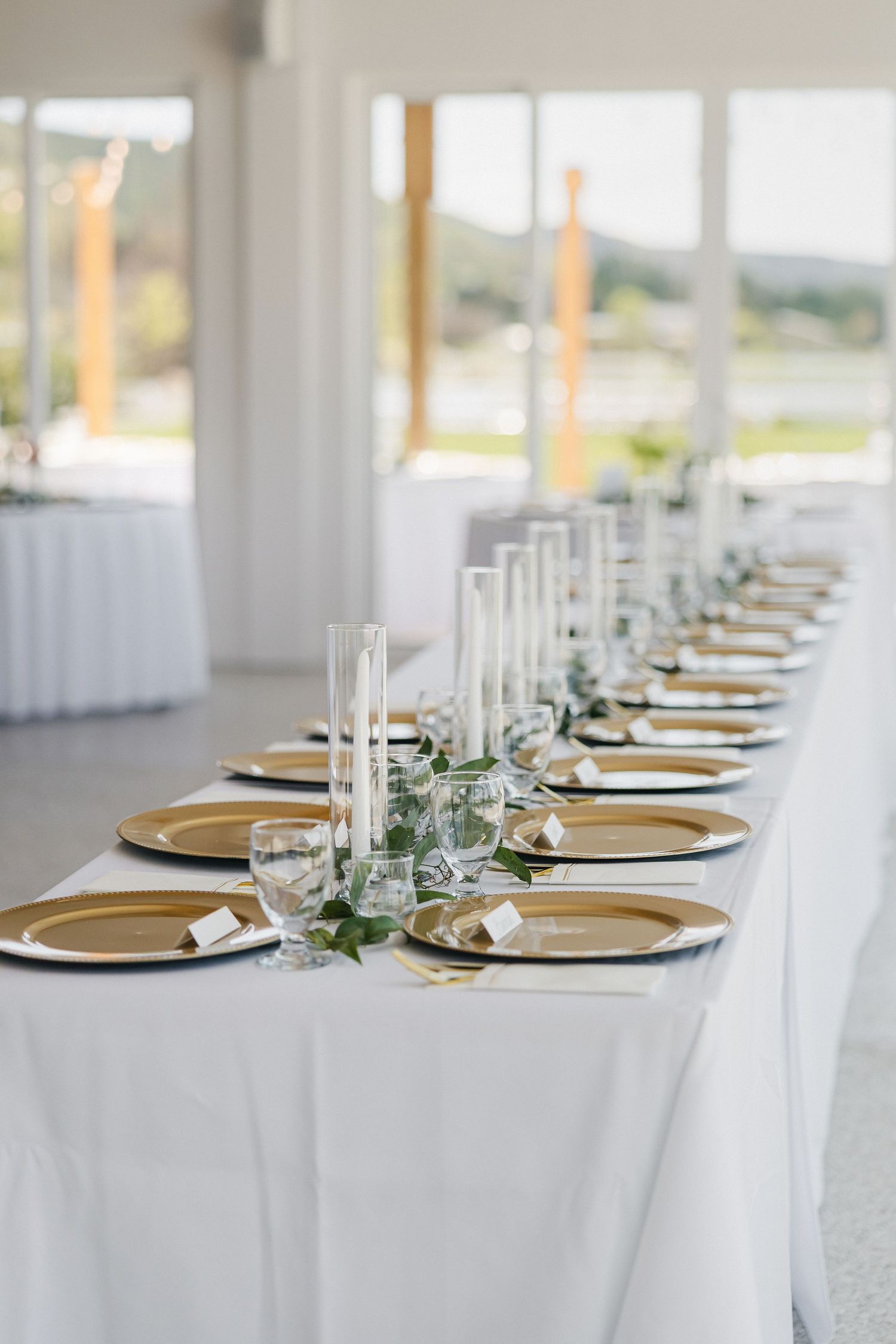Elegant wedding reception table with gold charger plates, white candles, and greenery on a white linen tablecloth.