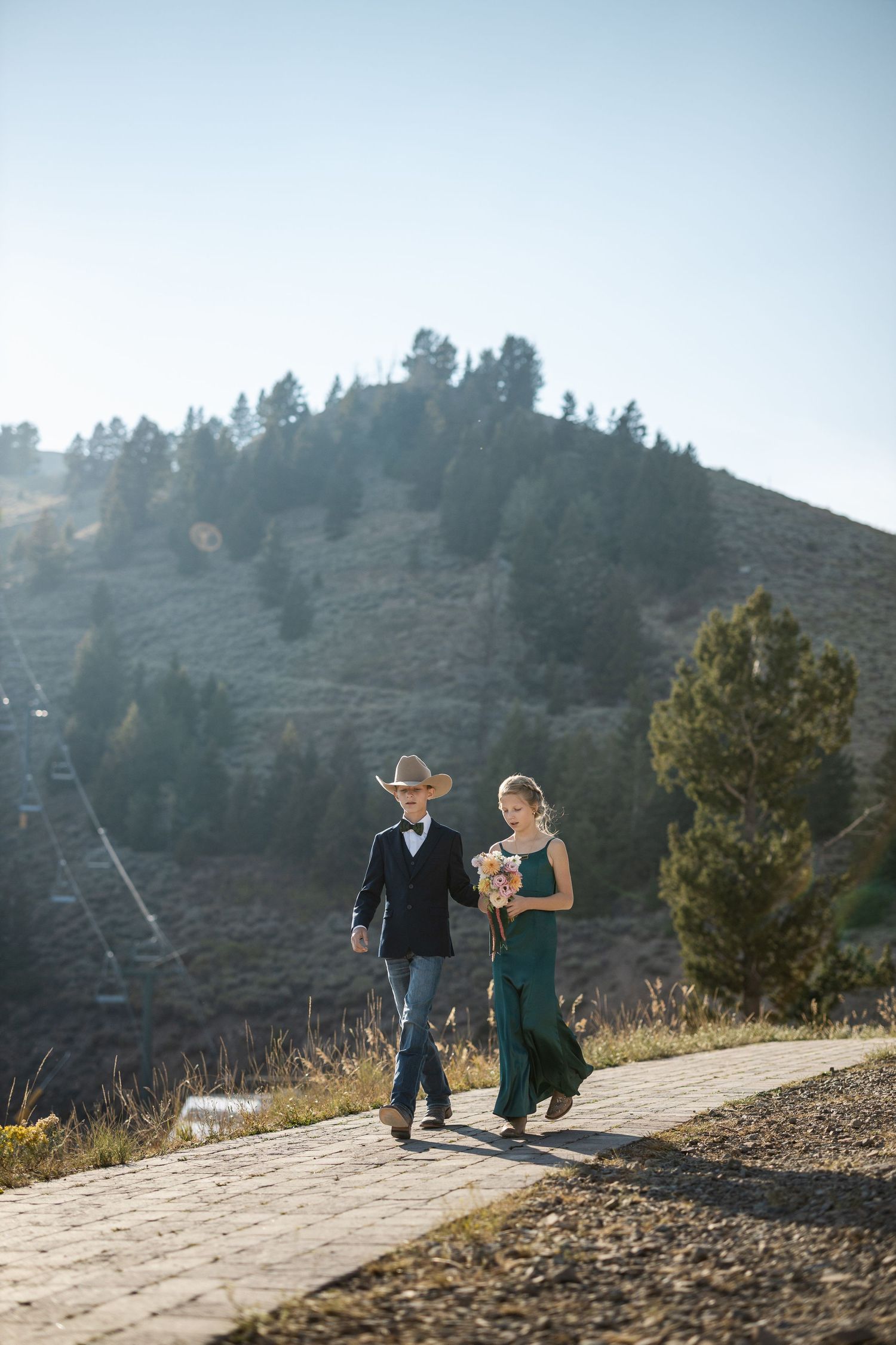 Couple strolls down a mountain path with pine trees and hills in the background.