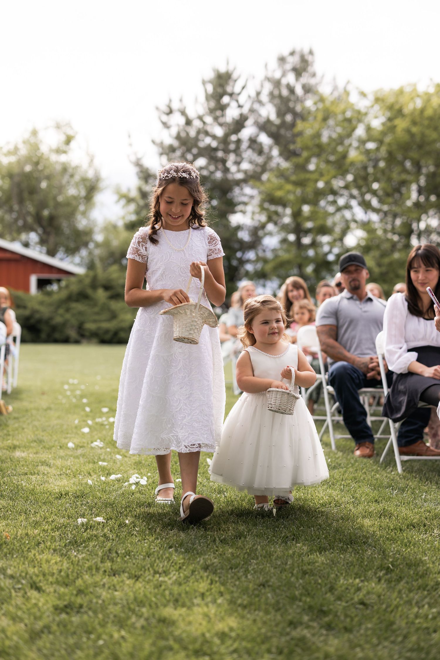 Young attendants in white dresses walk down a grassy aisle scattering flower petals.
