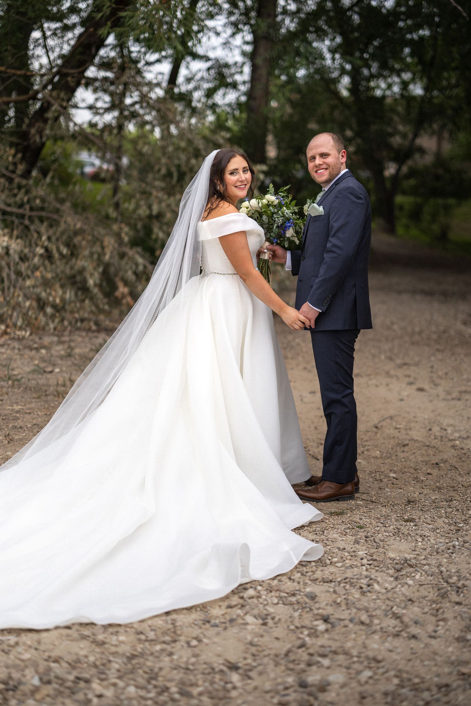 A wedding couple poses on a rustic path as the bride's flowing white dress trails behind them.