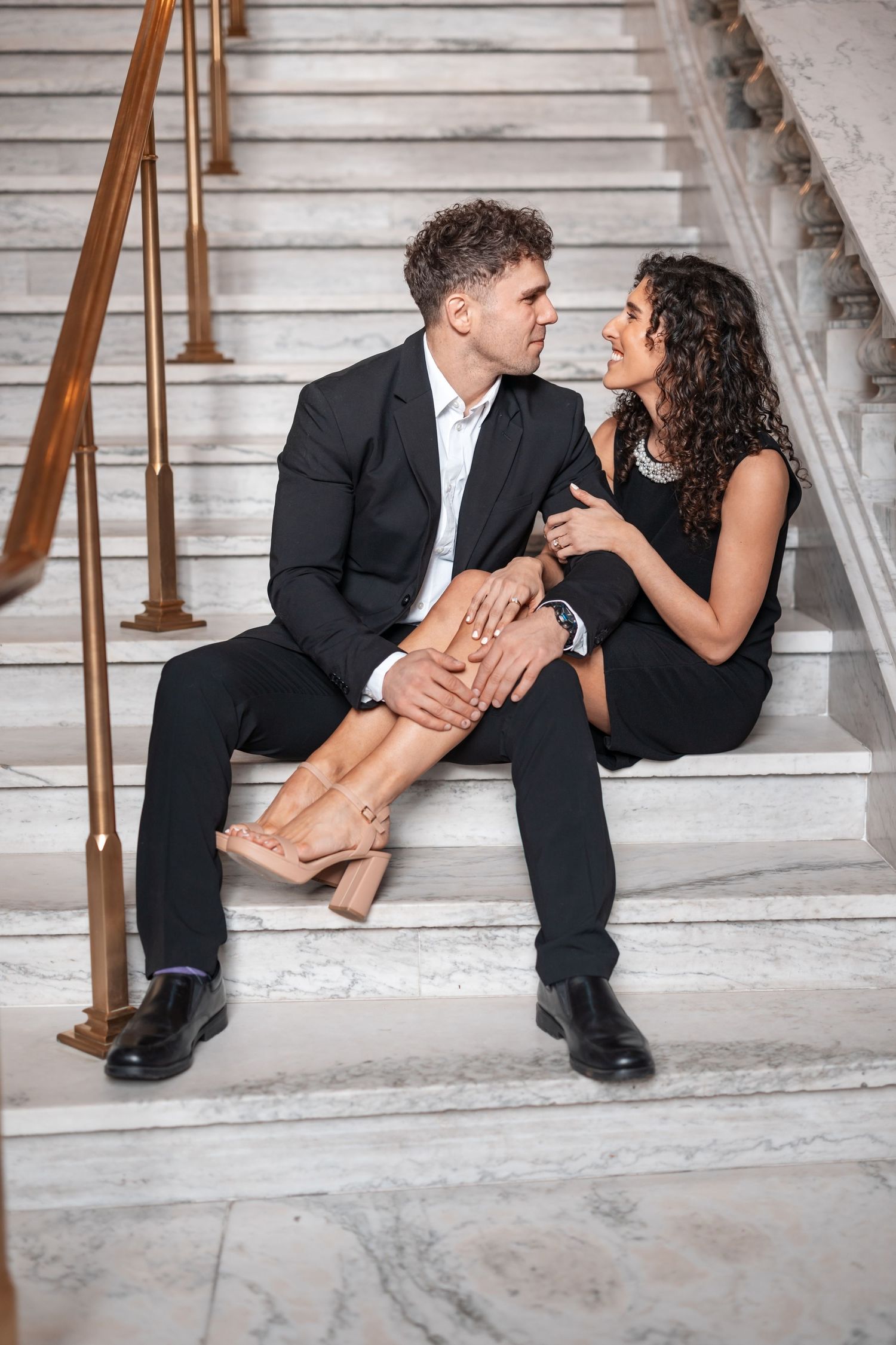 Couple in black and white attire shares tender moment on grand marble staircase.