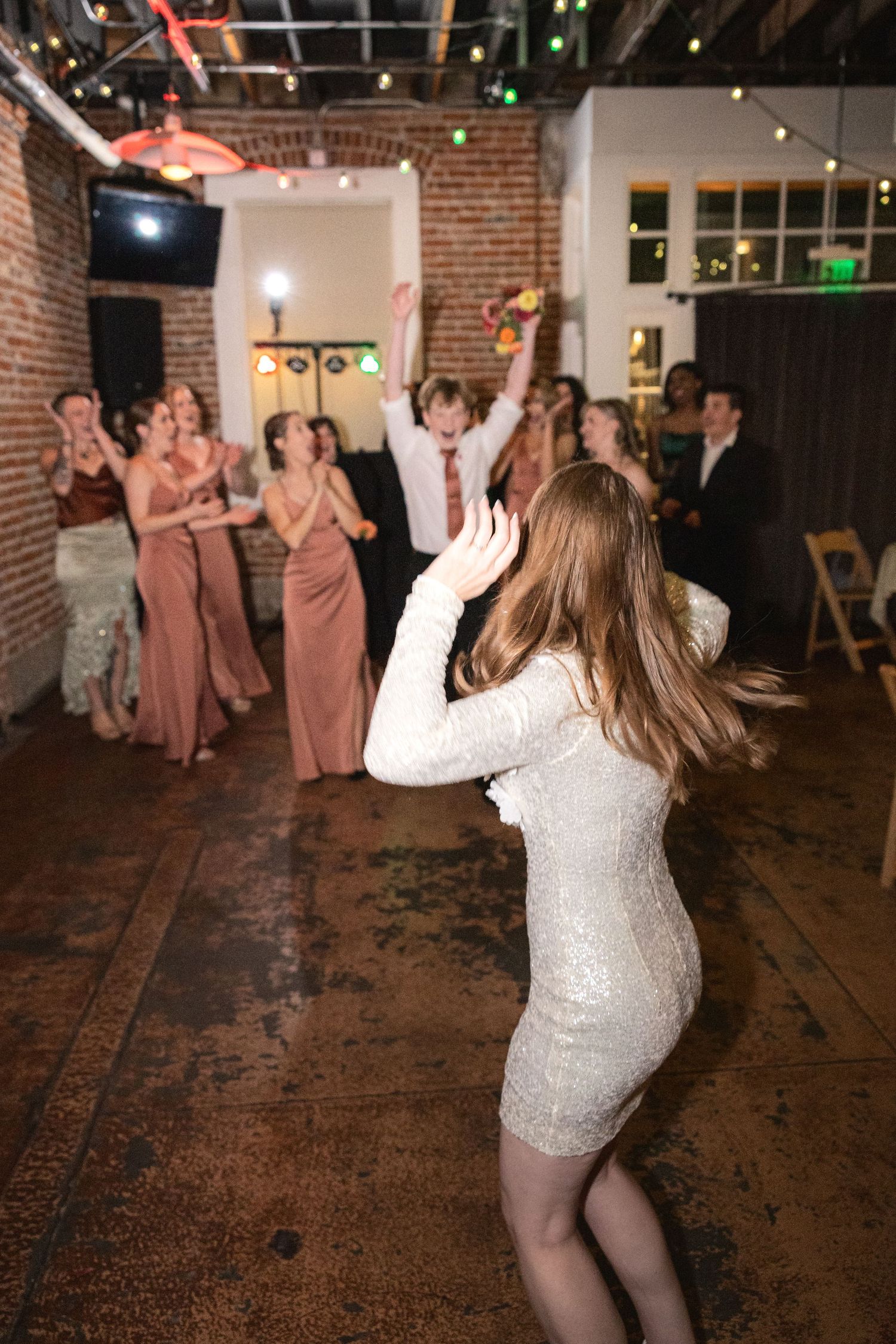 Wedding party members dance and celebrate on a rustic brick floor during a reception celebration.