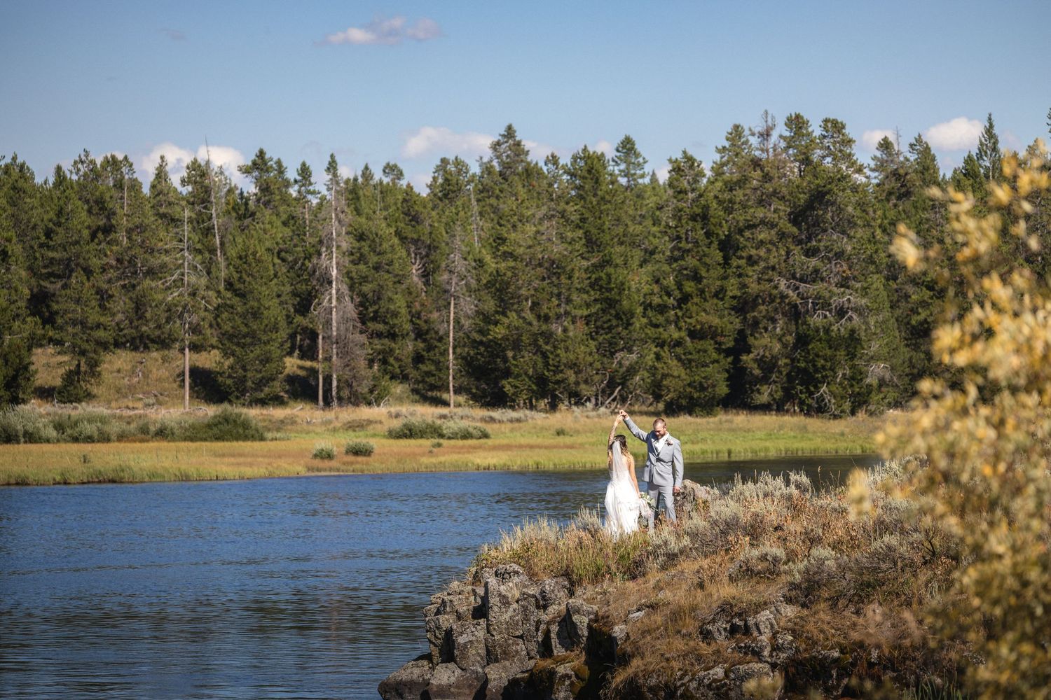 Couple in wedding attire pose on rocky cliff edge with forest and lake backdrop.