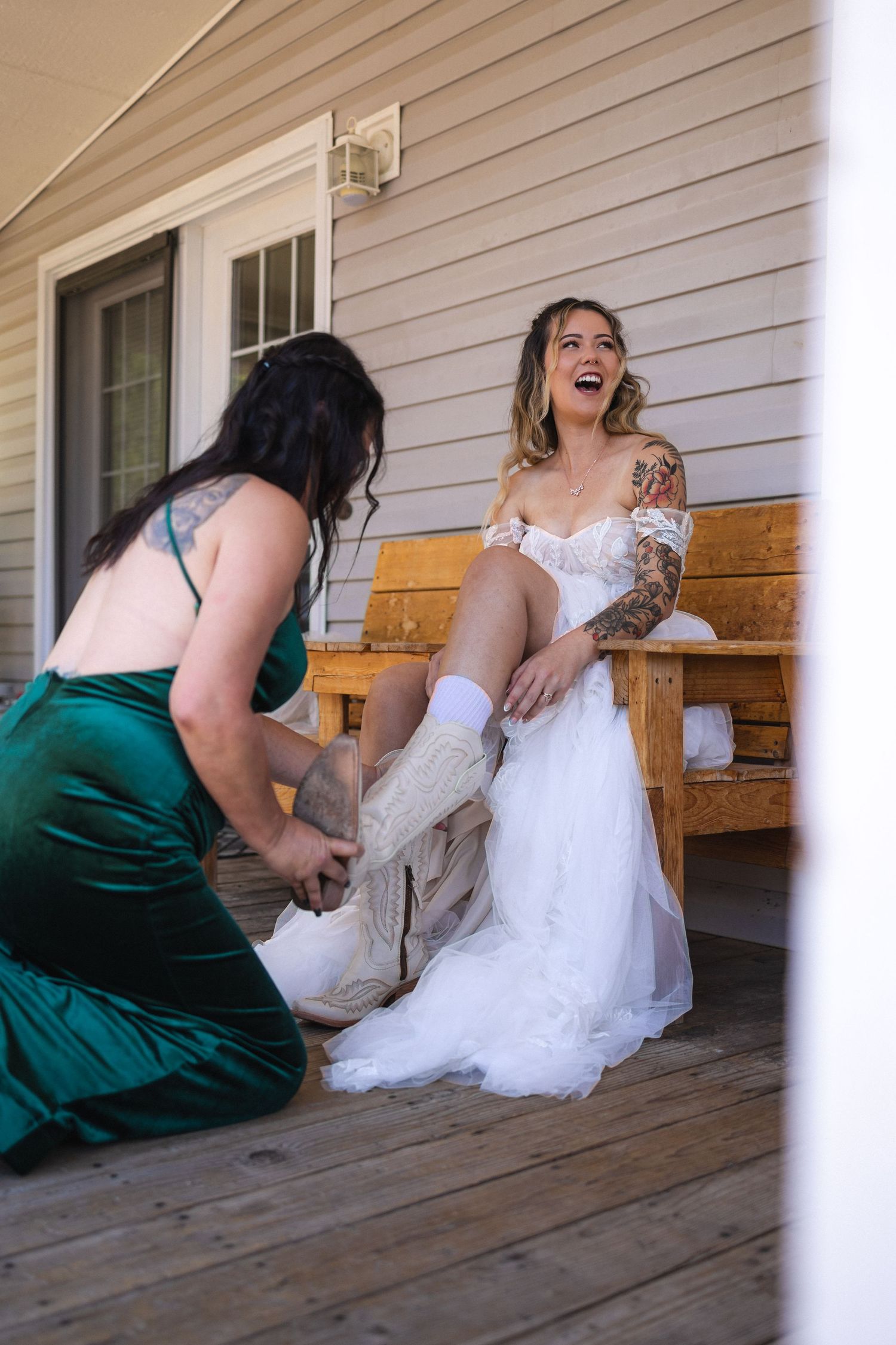 Joyful bride laughing while getting help putting on cowboy boots on rustic porch.
