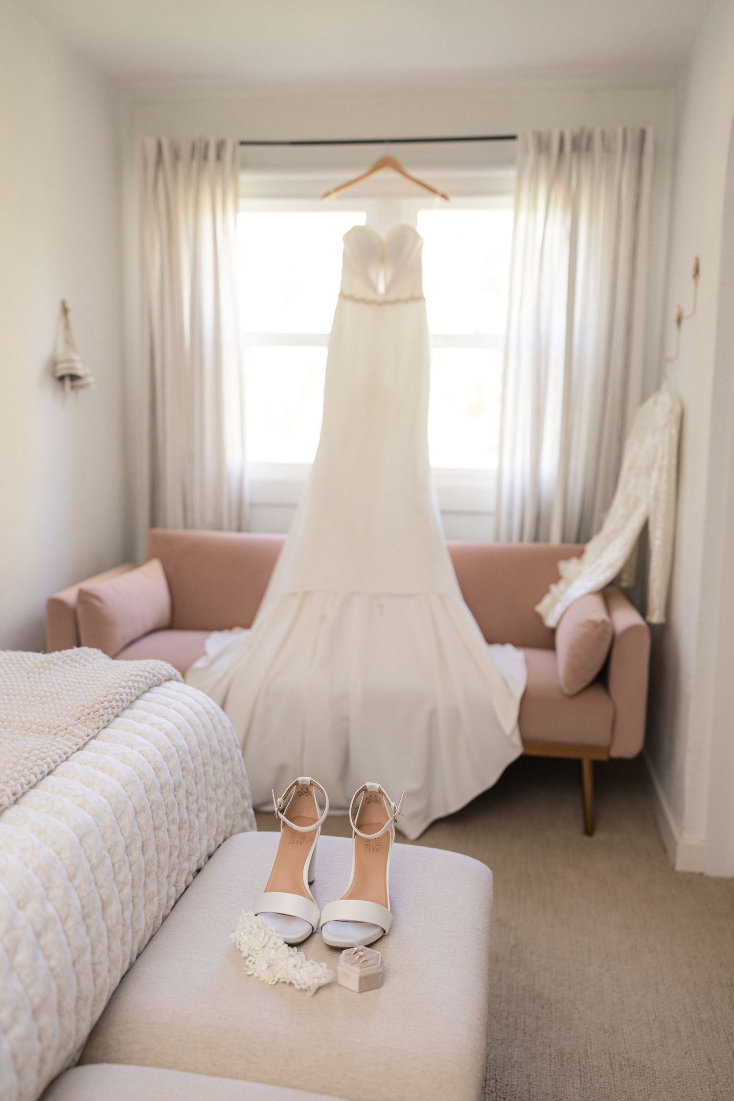 Elegant white wedding dress hangs in bright room with blush accents and white curtains.