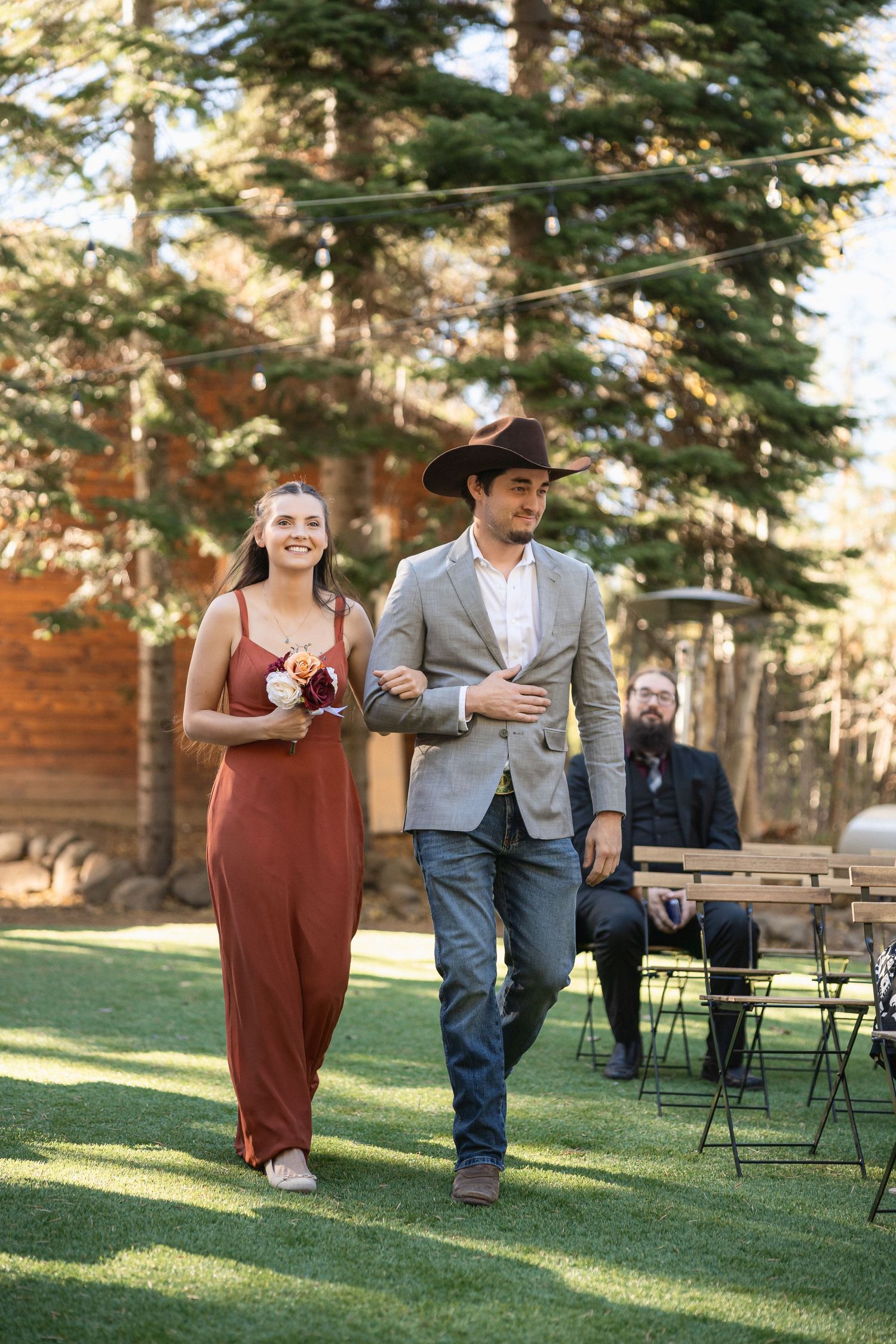 Attendants walking down outdoor wedding aisle with rustic decor and string lights.