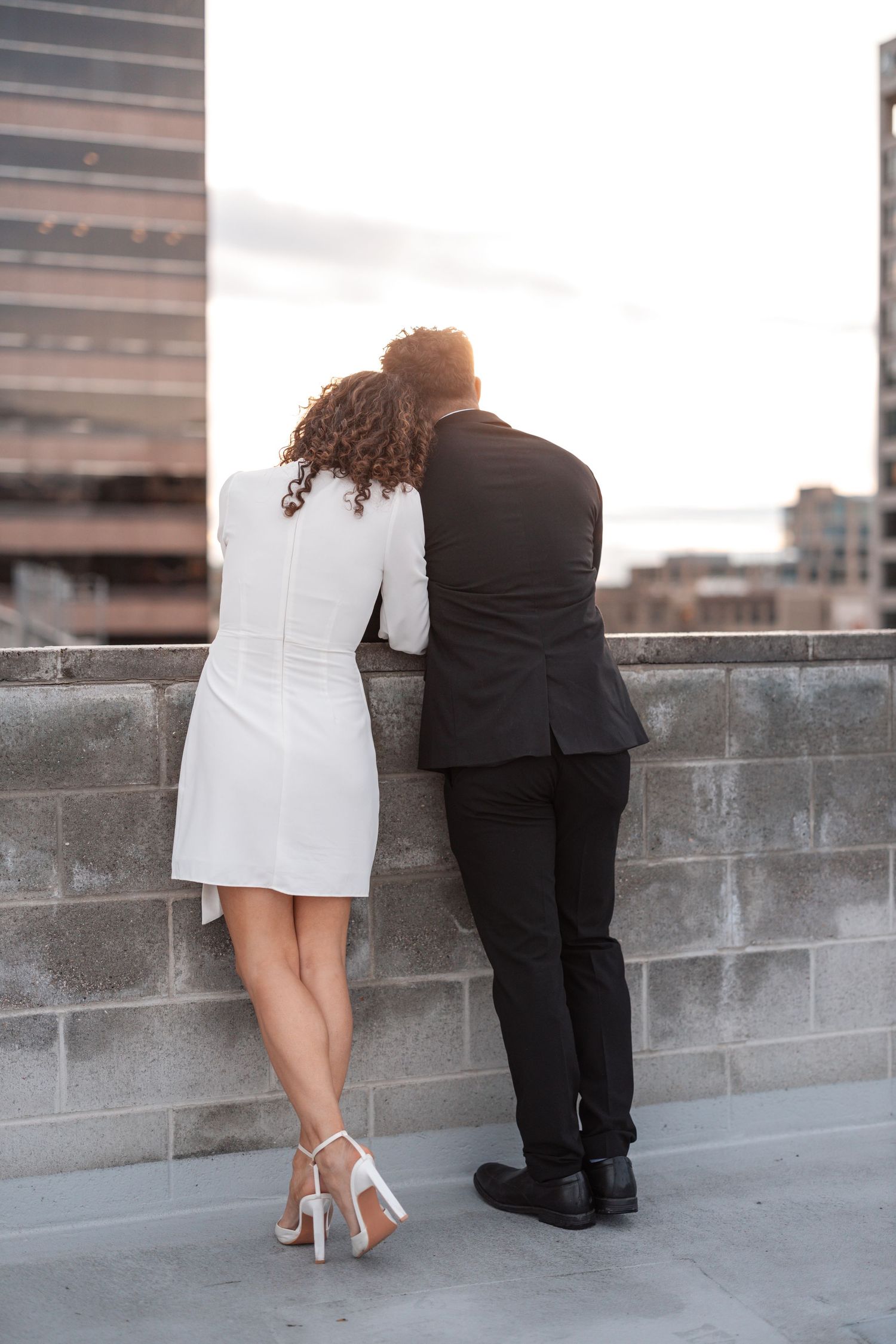 A couple in contrasting outfits stands at a rooftop wall overlooking the city.