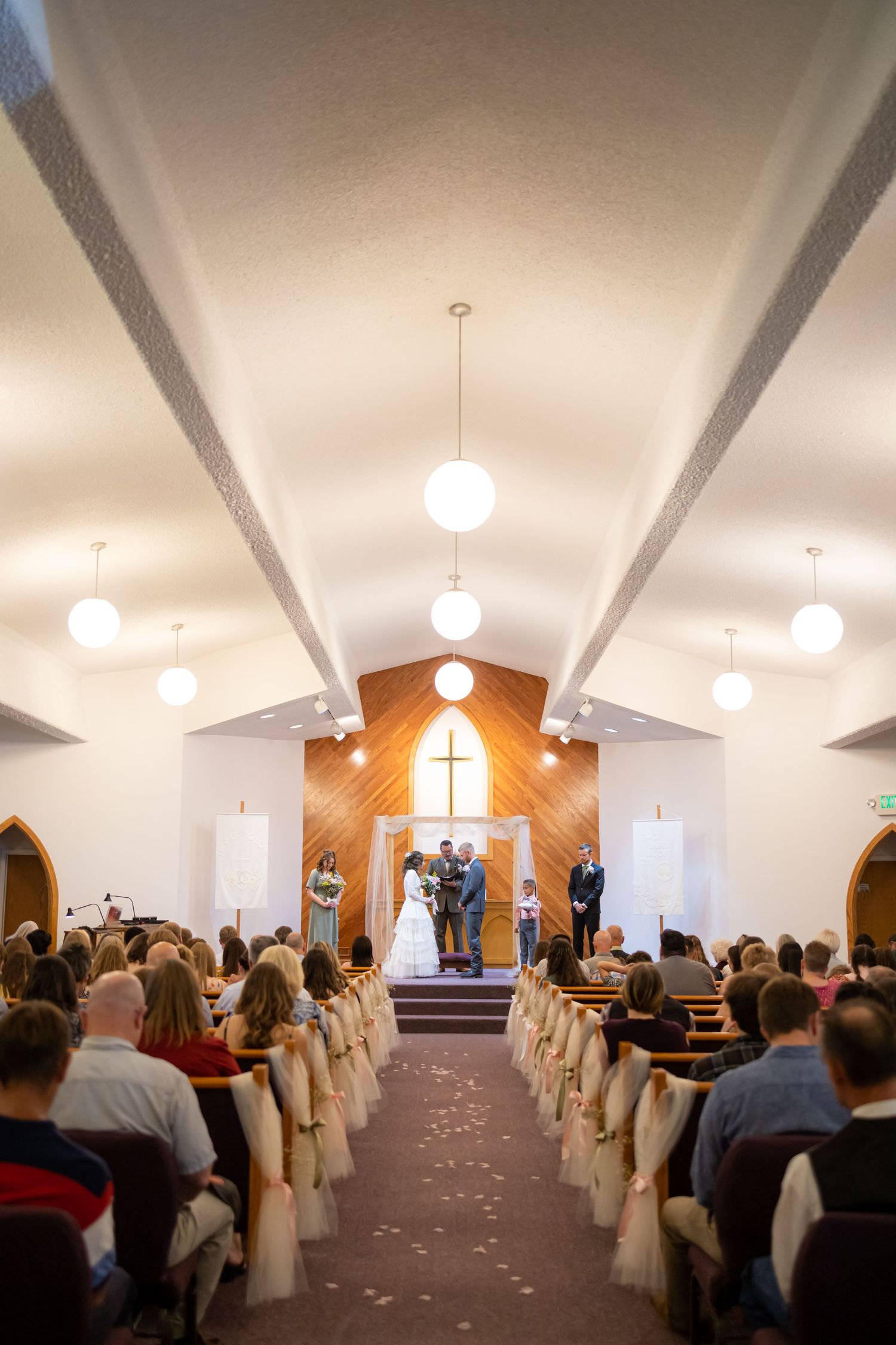 Church sanctuary filled with guests during a wedding ceremony with white chairs and globe lights.