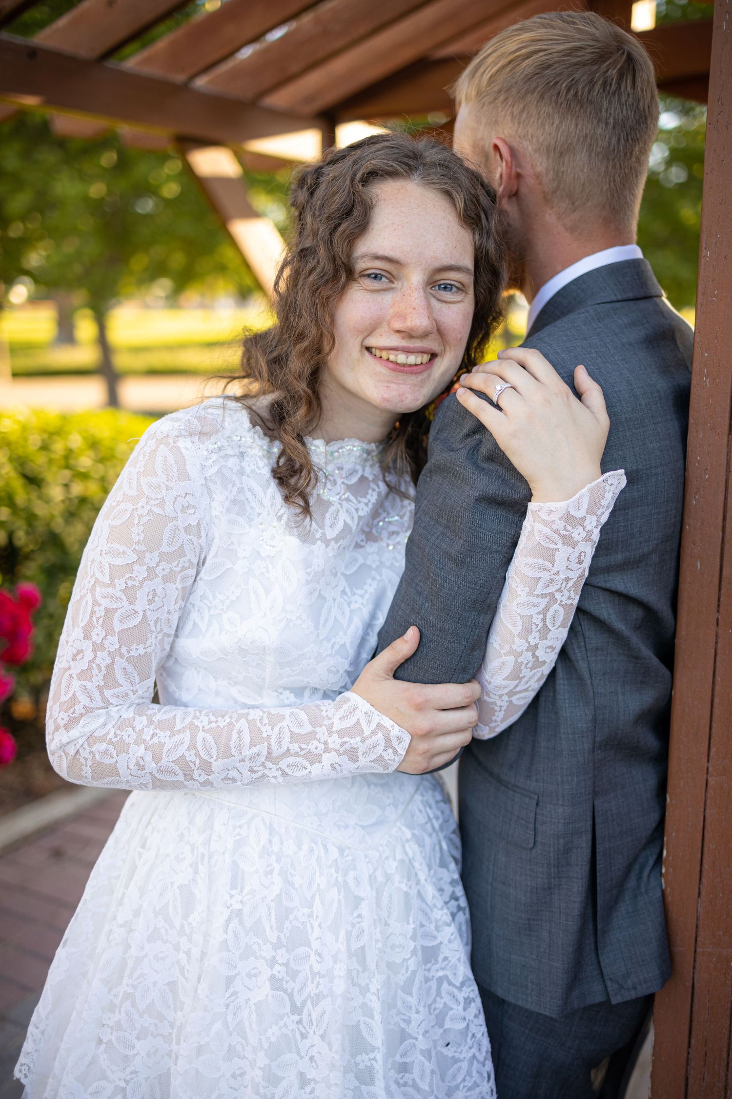 Intimate wedding portrait of couple embracing under wooden pergola surrounded by garden flowers.