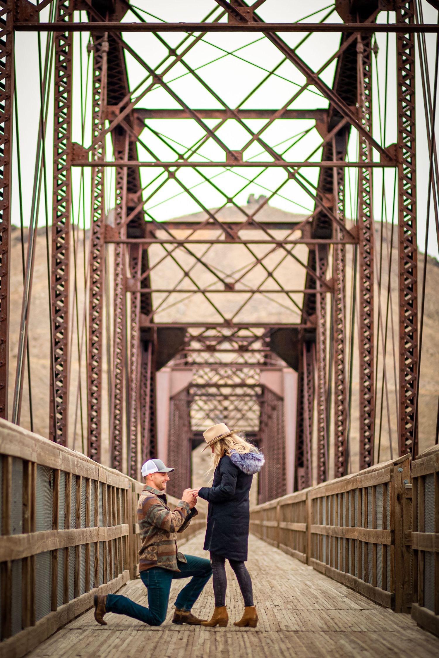 A romantic proposal moment on a rustic wooden bridge with metal truss architecture.